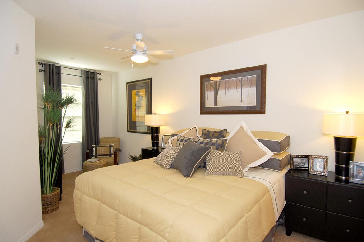 A cozy bedroom featuring a queen-sized bed with decorative pillows, two bedside tables with lamps, a small seating area with an armchair, and a framed artwork on the wall. Natural light streams in through a window covered by light curtains, complemented by indoor plants and a neutral color palette.