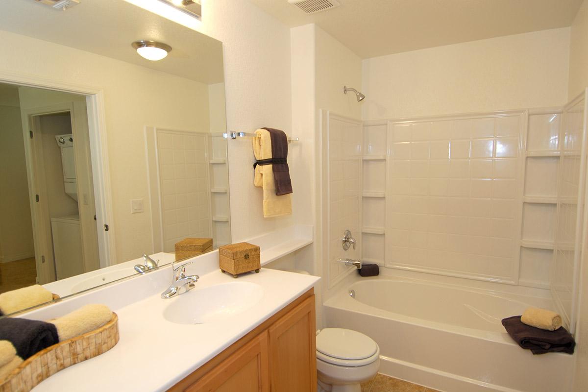 A clean and well-lit bathroom featuring a white bathtub with a shower, a sink with a mirror above it, and neatly arranged towels. The walls are a light color, and there are storage shelves in the bathtub area. A small basket sits on the countertop, adding a decorative touch.