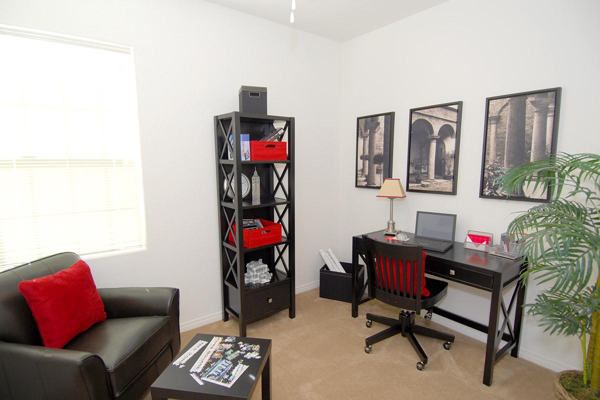 A cozy home office featuring a black leather armchair, a desk with a laptop and a red chair, a black bookshelf with red storage bins and decorative items, and framed wall art. There’s a green plant by the bookshelf and a window providing natural light. The room has neutral-colored walls and carpet.