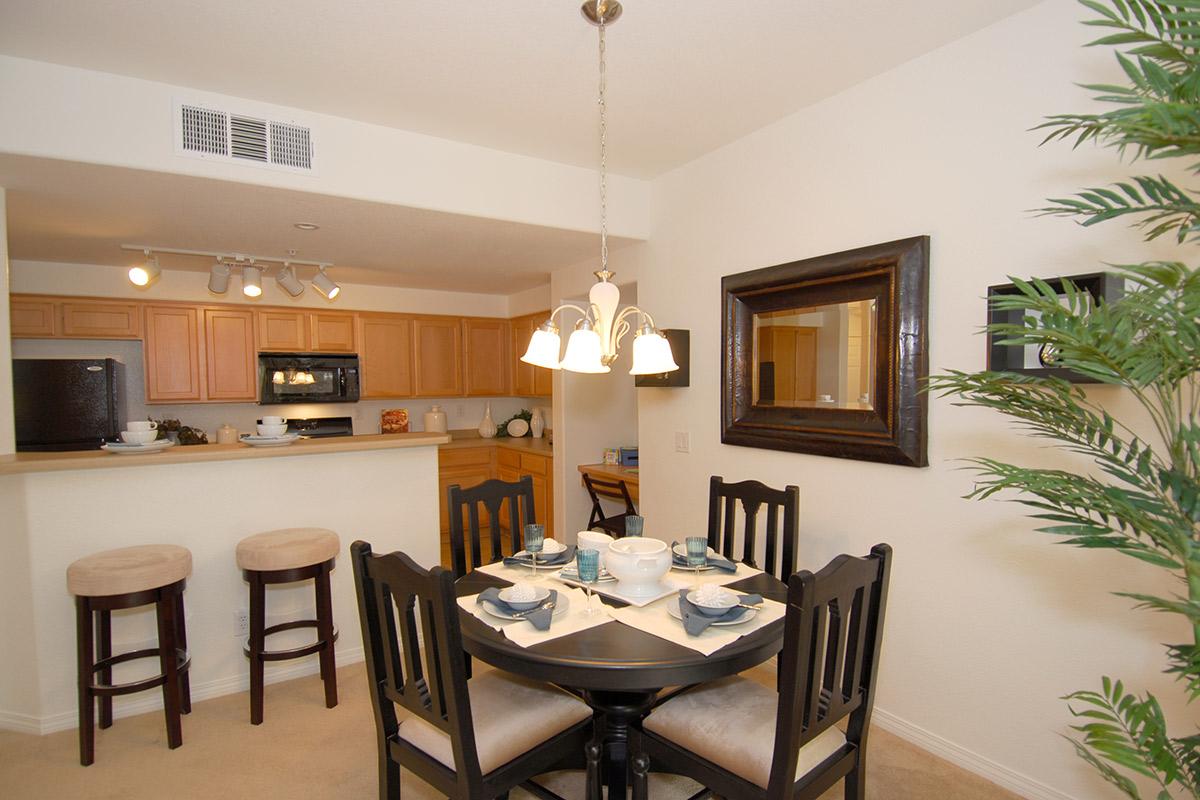 A cozy dining area featuring a round black table set for a meal with dishes and silverware. In the background, a modern kitchen with wooden cabinets, a dark countertop, and appliances. Two bar stools are positioned near the kitchen, and a decorative plant adds a touch of greenery to the space.