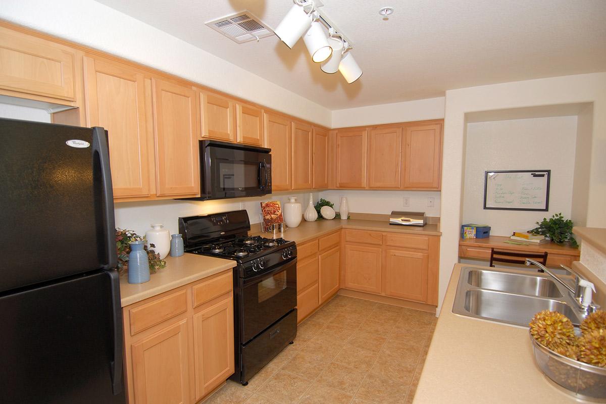 Modern kitchen featuring light wooden cabinetry, black appliances including a stove and refrigerator, and a single basin sink. The countertops are beige, and decorative elements like plants and vases are visible. There is a whiteboard on the wall and a warm, inviting atmosphere.