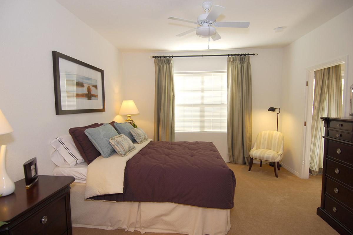 A cozy bedroom featuring a queen-sized bed with a dark purple duvet and decorative pillows. A nightstand with a lamp and framed artwork adorn the walls. Light-colored curtains frame a window, and a small, stylish armchair is positioned beside a dresser, complementing the warm, inviting atmosphere.