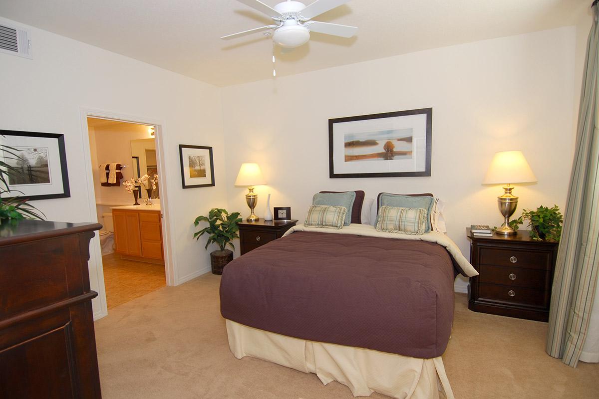 A cozy bedroom featuring a queen-sized bed with a brown quilt, two bedside lamps, and artwork on the wall. There's a dresser and a potted plant, with a doorway leading to a bathroom in the background. The room has warm lighting and neutral-toned walls.