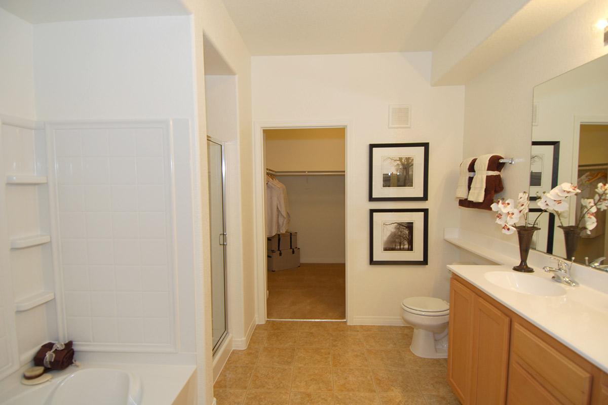 A bright bathroom featuring a white tub, glass shower, and beige tile floor. The vanity has wooden cabinets with a sink and a large mirror. On the wall, there are two framed black-and-white photos, and a small vase with artificial flowers. An open closet with hangers is visible in the background.