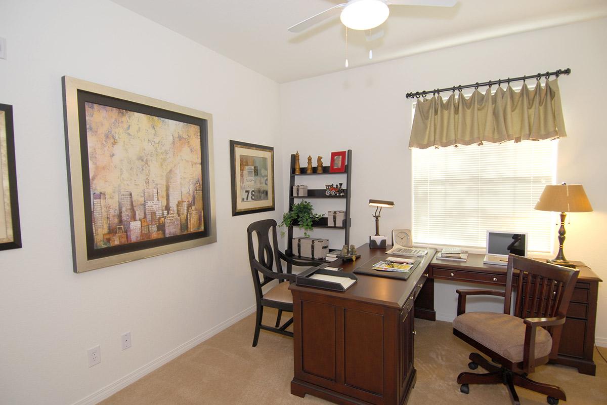 A well-organized office space featuring a wooden desk with a chair, books, and a lamp. The wall is adorned with framed artwork and photographs. A window with a curtain allows natural light to brighten the room. The decor includes decorative items on shelves and a comfortable seating area.