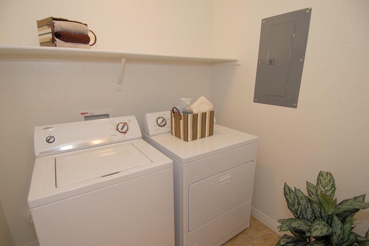 A laundry room featuring a washing machine and a dryer side by side. Above the machines, there is a shelf holding decorative items. A storage basket with toiletries is placed on the dryer. A small potted plant is positioned on the floor, and an electrical panel is mounted on the wall.