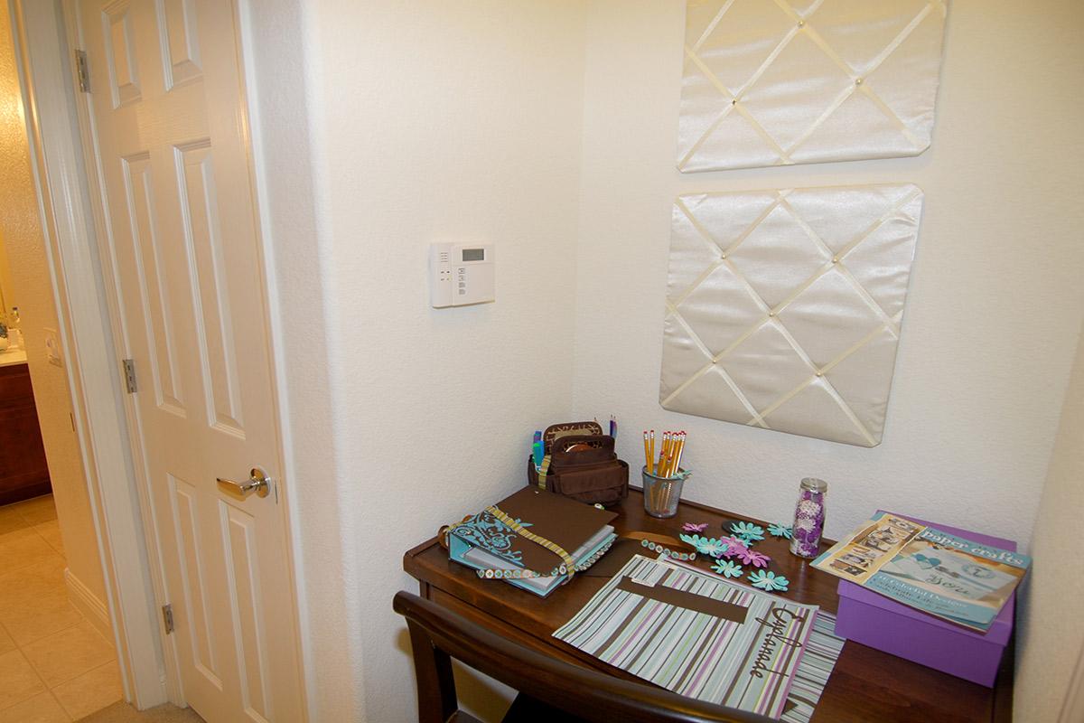 A corner of a room featuring a wooden desk with notebooks, pens, and decorative items. Two quilted wall panels are displayed above the desk. A control panel is mounted on the wall, and a bathroom entrance is visible in the background. The floor is tiled, and the walls are painted a light color.
