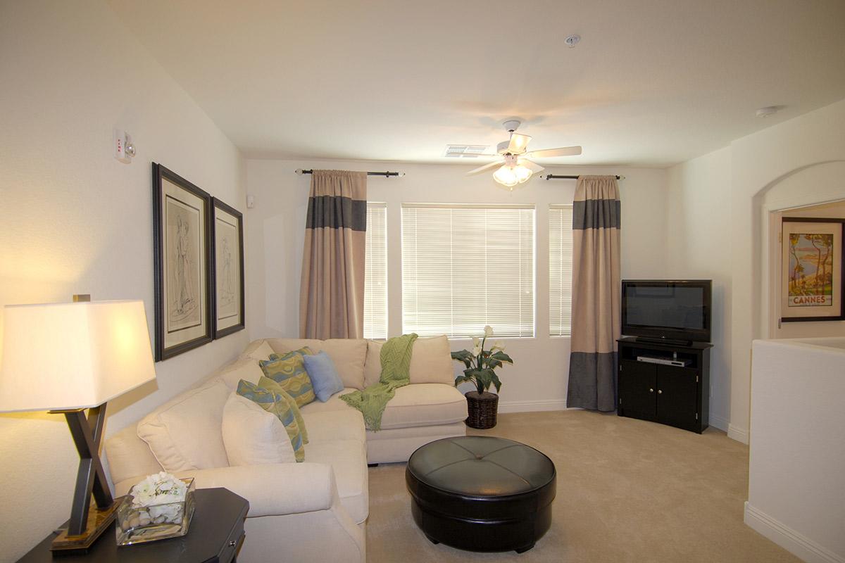 A cozy living room with a light-colored sectional sofa adorned with colorful cushions. There is a round coffee table in front of the sofa, a potted plant, and a television against the wall. Large windows with curtains allow natural light to enter, creating a warm and inviting atmosphere.