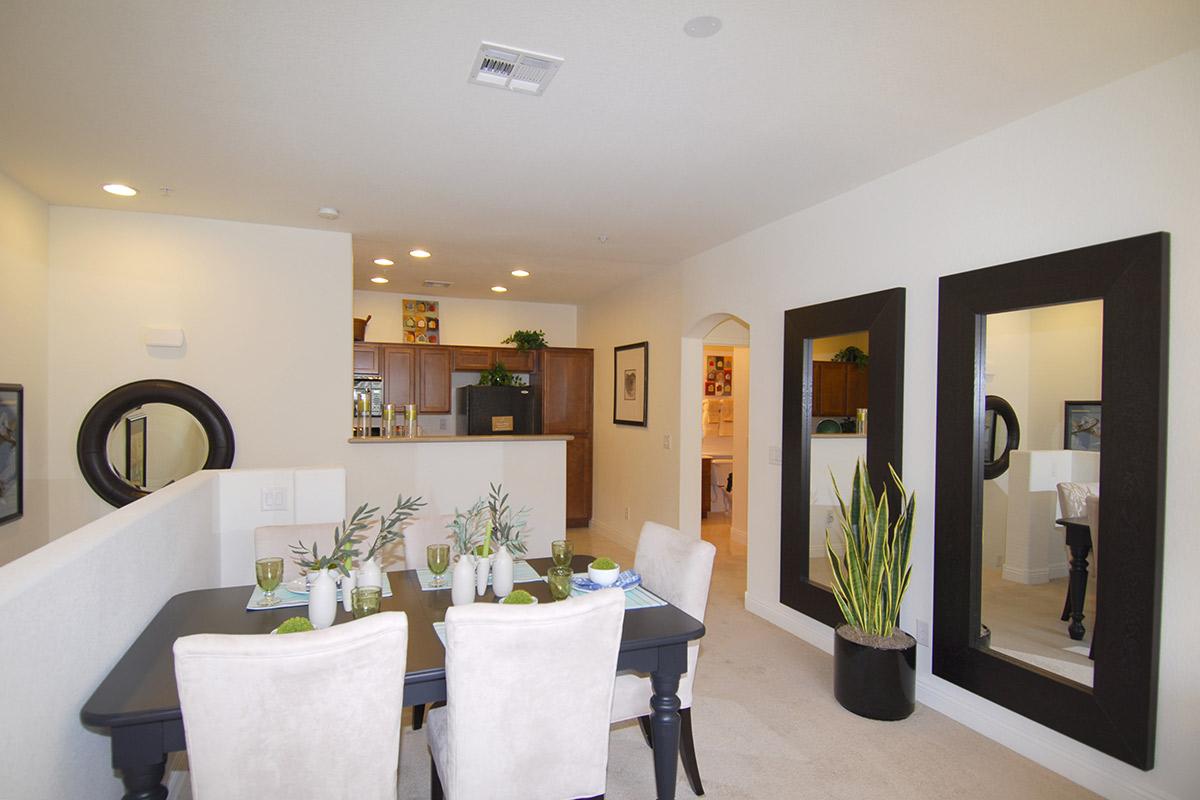 A modern dining area featuring a black dining table set with decorative plants and green tableware. The space includes two large mirrors on the wall, warm lighting, and a view of a kitchen with wooden cabinetry in the background. The overall decor is light and inviting.