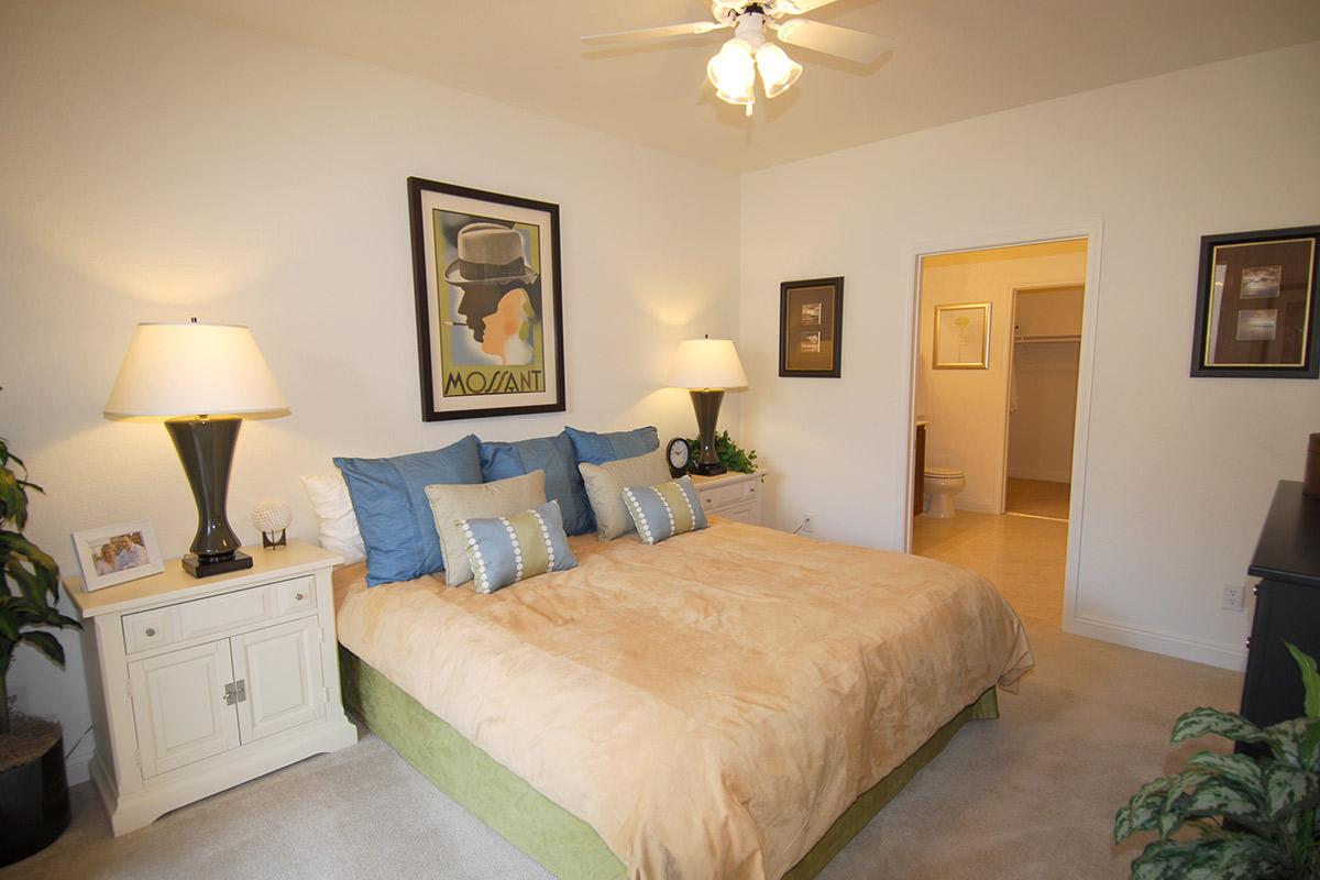 A well-decorated bedroom featuring a large bed with beige and blue pillows, matching lamps on either side, and a stylish poster on the wall. The room has a warm, inviting atmosphere with light-colored walls and a soft carpet, along with a small plant and a door leading to a bathroom.