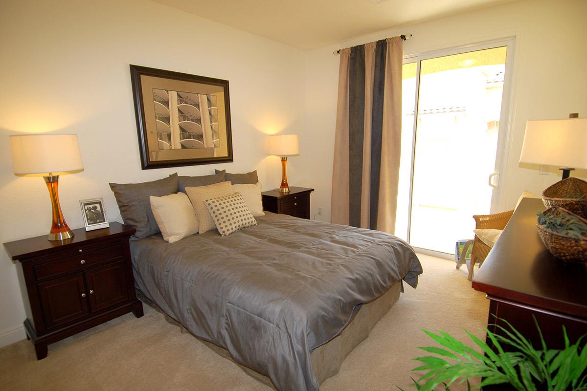 Cozy bedroom featuring a queen-sized bed with gray bedding and decorative pillows. Two bedside tables with lamps are placed on either side, and there is a framed artwork above the bed. A large window with curtains lets in natural light, complementing the neutral color scheme and creating a warm atmosphere.