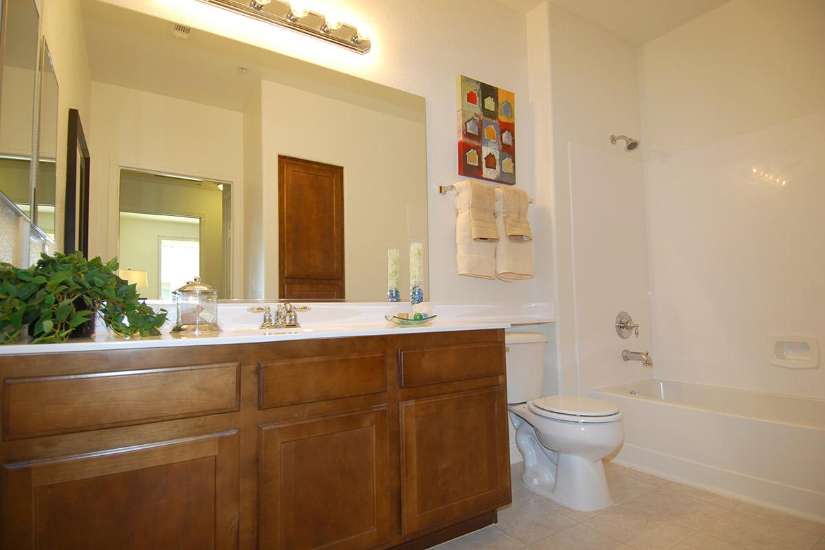 A bright and clean bathroom featuring a wooden vanity with double sinks, a large mirror, and a bathtub with a shower. The walls are painted light, and decorative items include candles and a small plant. A colorful piece of artwork is mounted above the sink area, adding a splash of color to the space.