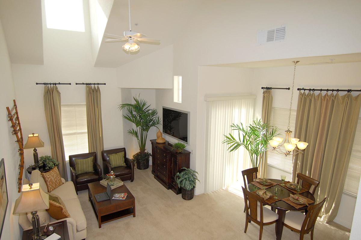 An aerial view of a bright and spacious living area featuring a sofa, two armchairs, and a dining table set for four. Large windows with drapes allow natural light to fill the room, complemented by indoor plants and warm decor, creating a cozy and inviting atmosphere.