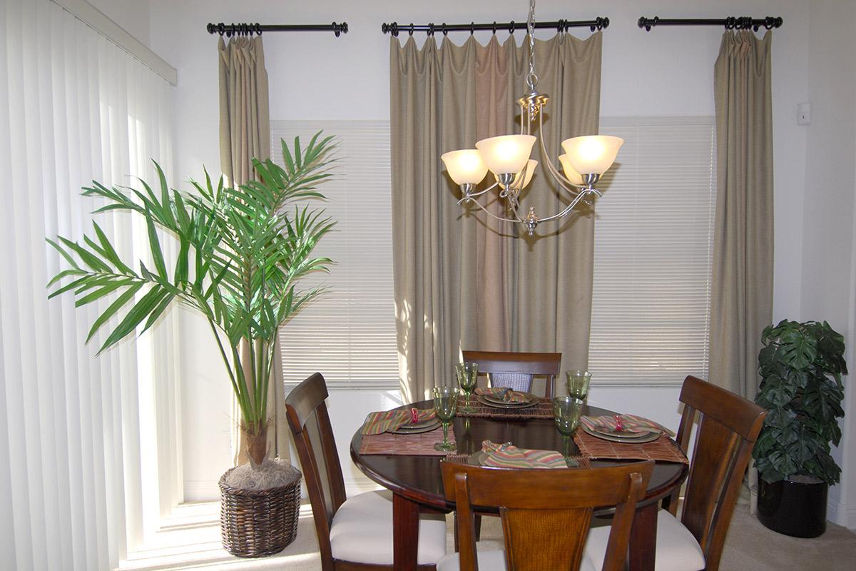 A well-lit dining area featuring a round wooden table set for four with green glassware and patterned placemats. A large potted palm and a smaller leafy plant are visible, along with elegant curtains draping the windows, creating a warm and inviting atmosphere.