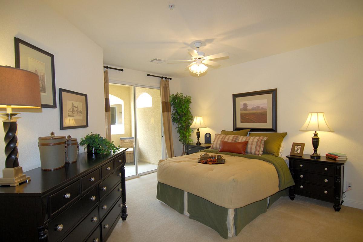 Cozy bedroom featuring a neutral color scheme with a queen-sized bed adorned in green and beige bedding. Two nightstands with lamps flank the bed, and framed artwork is displayed on the walls. A ceiling fan provides additional comfort, while a sliding door leads to a patio area.