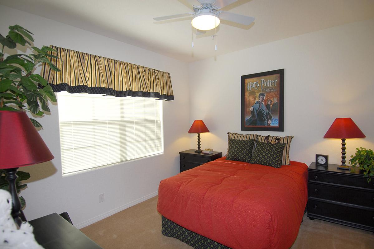 Cozy bedroom featuring a red bedspread, black nightstands with red lamps, and a large poster of a character from Harry Potter on the wall. The window has a striped valance, and there is a potted plant in the corner, creating a warm and inviting atmosphere.