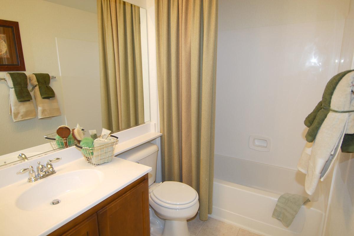 A small bathroom featuring a white bathtub and shower with a shower curtain. A sink with a wooden cabinet below, a mirror, and various toiletries are visible. There are green towels hanging on the wall and a decorative basket on the counter. The room has light-colored walls and a cozy, neutral decor.