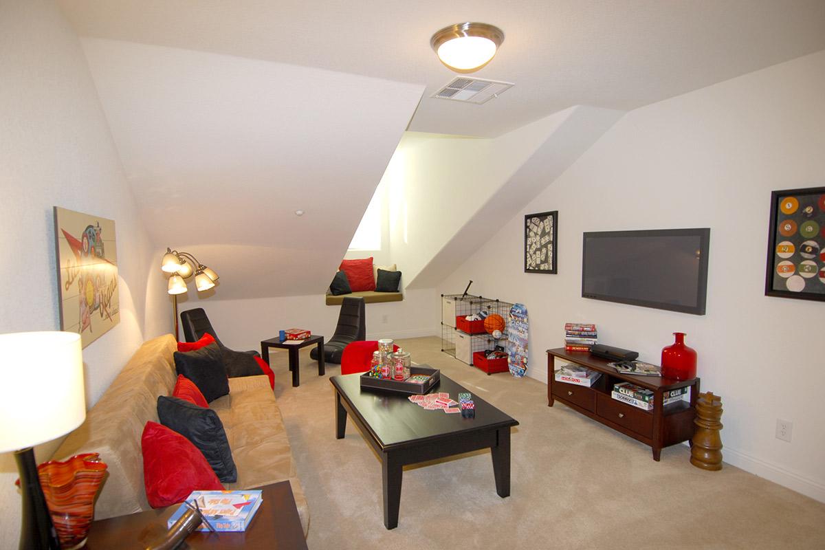 Cozy, modern living room featuring a beige couch with red and black throw pillows, two black chairs, a coffee table with decorations, and a flat-screen TV. The walls are light-colored, and there are decorative items, including artwork and games, on display. Soft lighting creates a warm atmosphere.