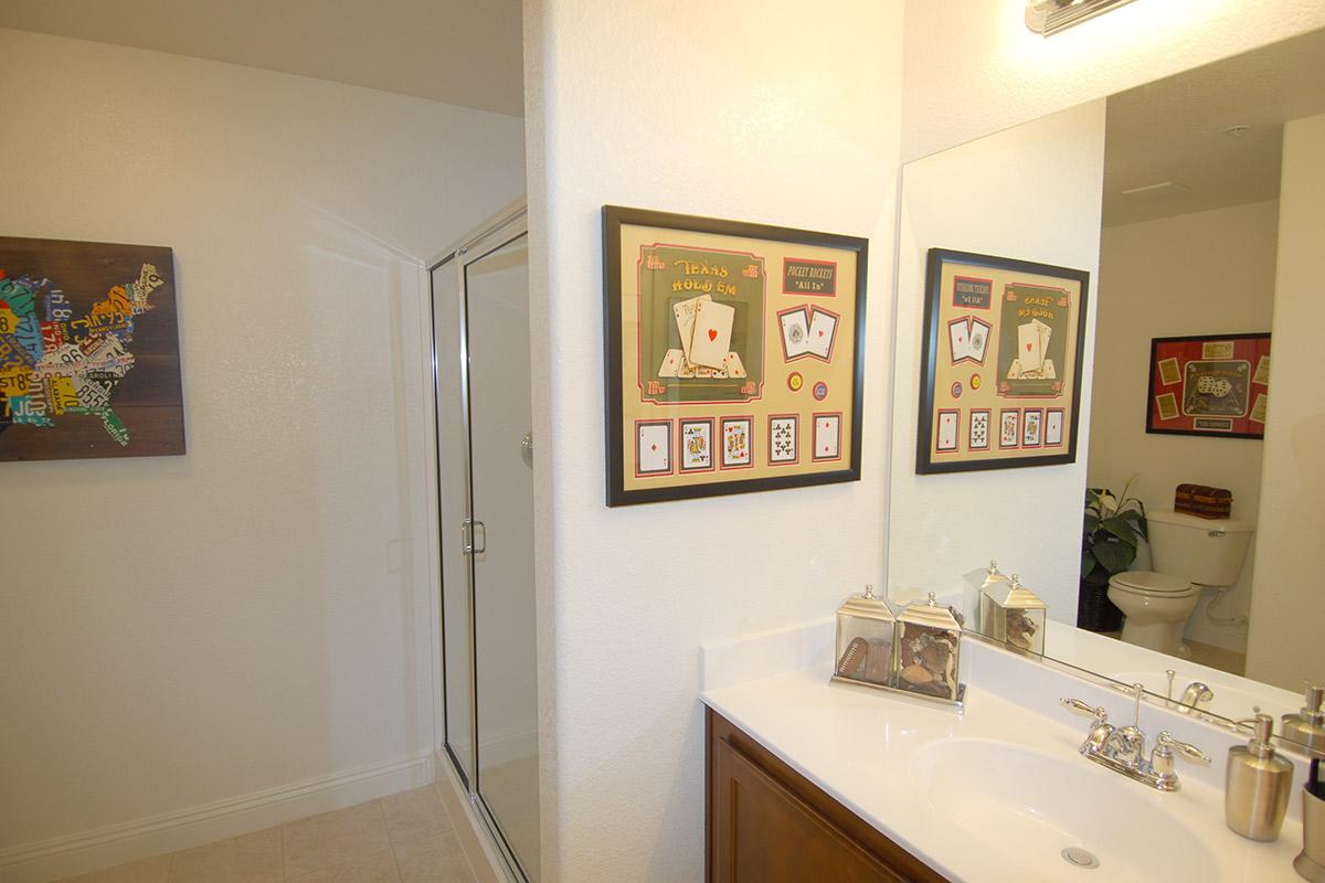 A bright bathroom featuring a large mirror above a white sink with wooden cabinetry. On the wall, there are two framed art pieces related to Texas hold 'em poker, alongside a decorative box. A sliding glass shower door is visible, and a toilet can be seen in the background, with light-colored walls and tiles.