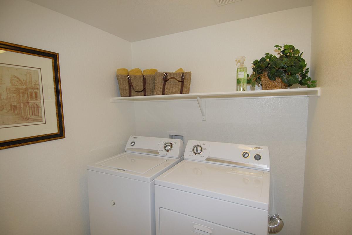 A clean laundry room featuring a white washing machine and dryer side by side. Above them, a shelf holds two baskets and decorative items, including a plant and cleaning supplies. A framed artwork hangs on the wall, adding a stylish touch to the space.