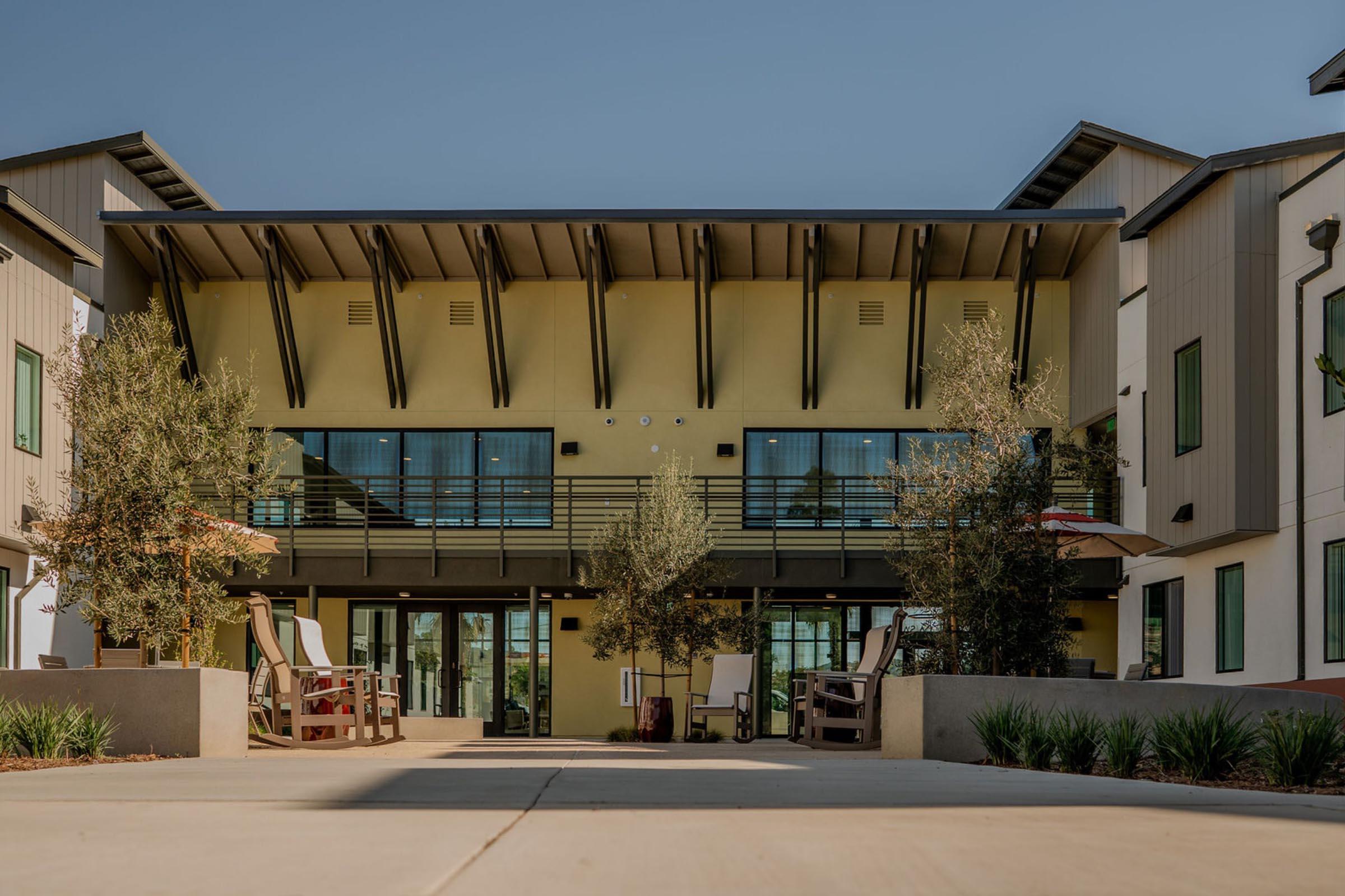 Modern multi-story building exterior featuring large windows, a balcony, and a welcoming entrance. Outdoor seating areas with lounge chairs and umbrellas are positioned in front, surrounded by landscaped greenery and pathways, under a clear blue sky.
