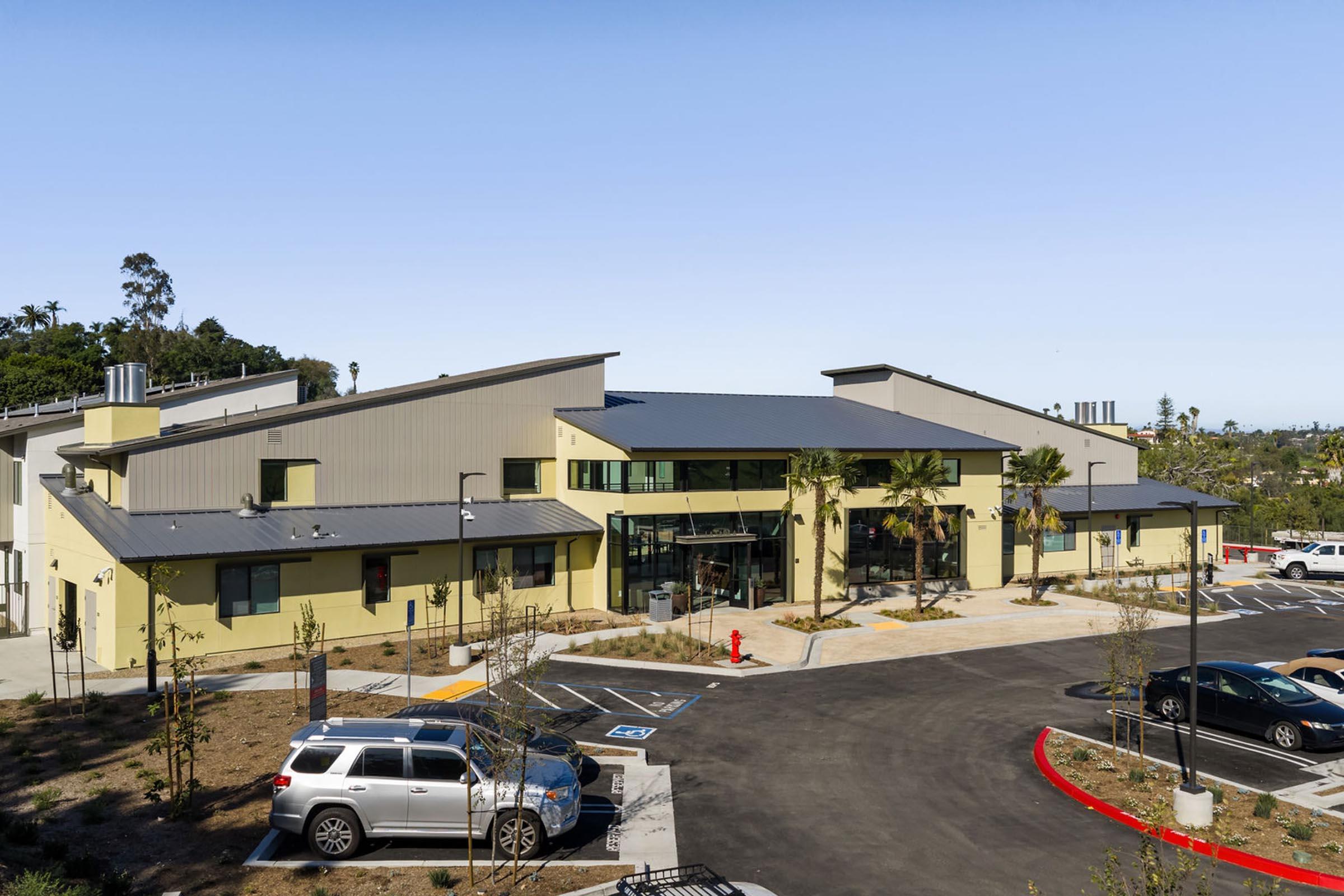 A modern building with a sloped roof, featuring large windows and a welcoming entrance. The structure is surrounded by landscaped areas and parking spaces. Palm trees are planted nearby, and the setting is sunny with clear skies.