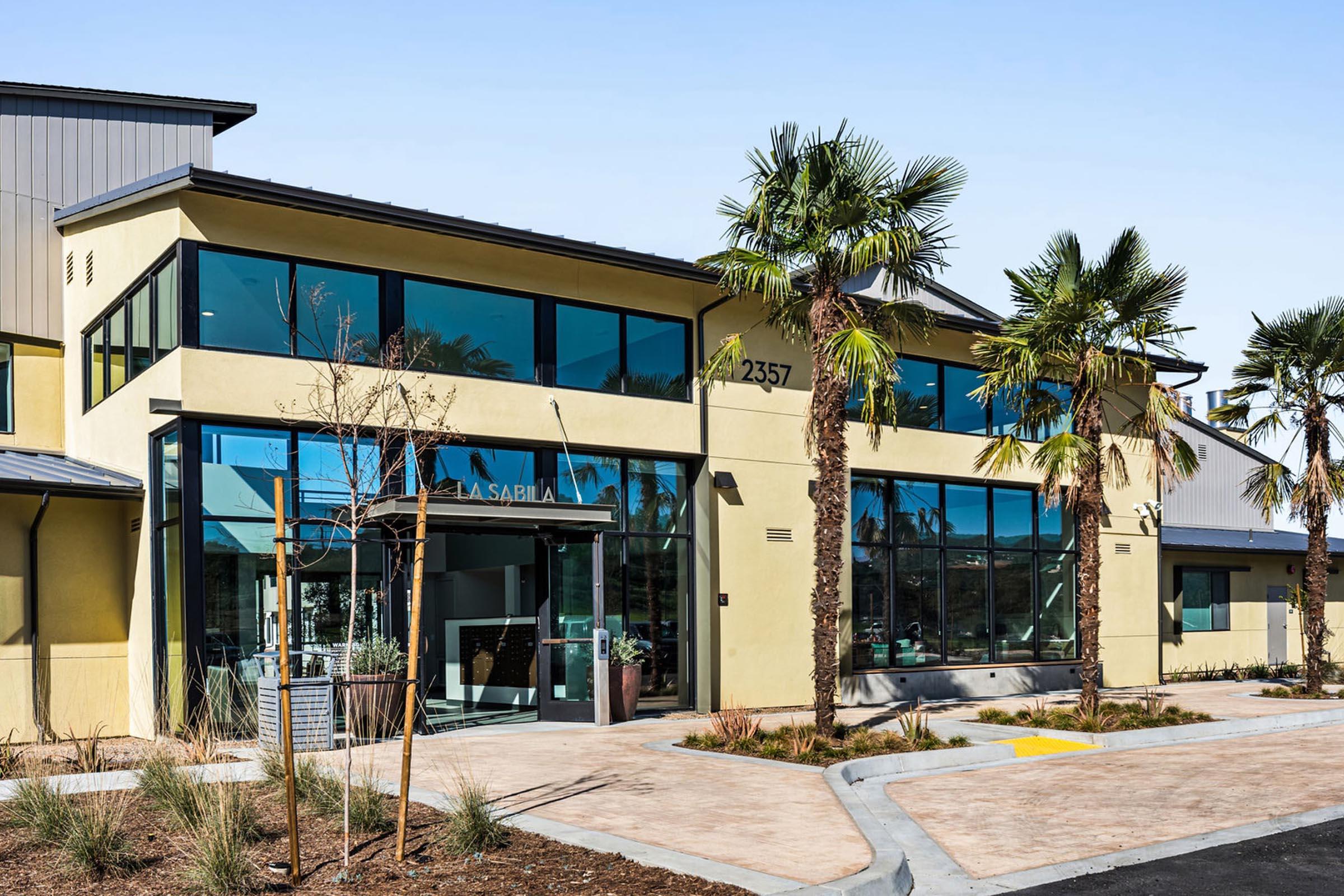 Modern building exterior featuring large windows and palm trees. The structure has a light yellow facade and is designed with a mix of contemporary elements. The entrance is prominently displayed, showcasing a welcoming atmosphere. Landscaping includes paved walkways and sparse greenery.