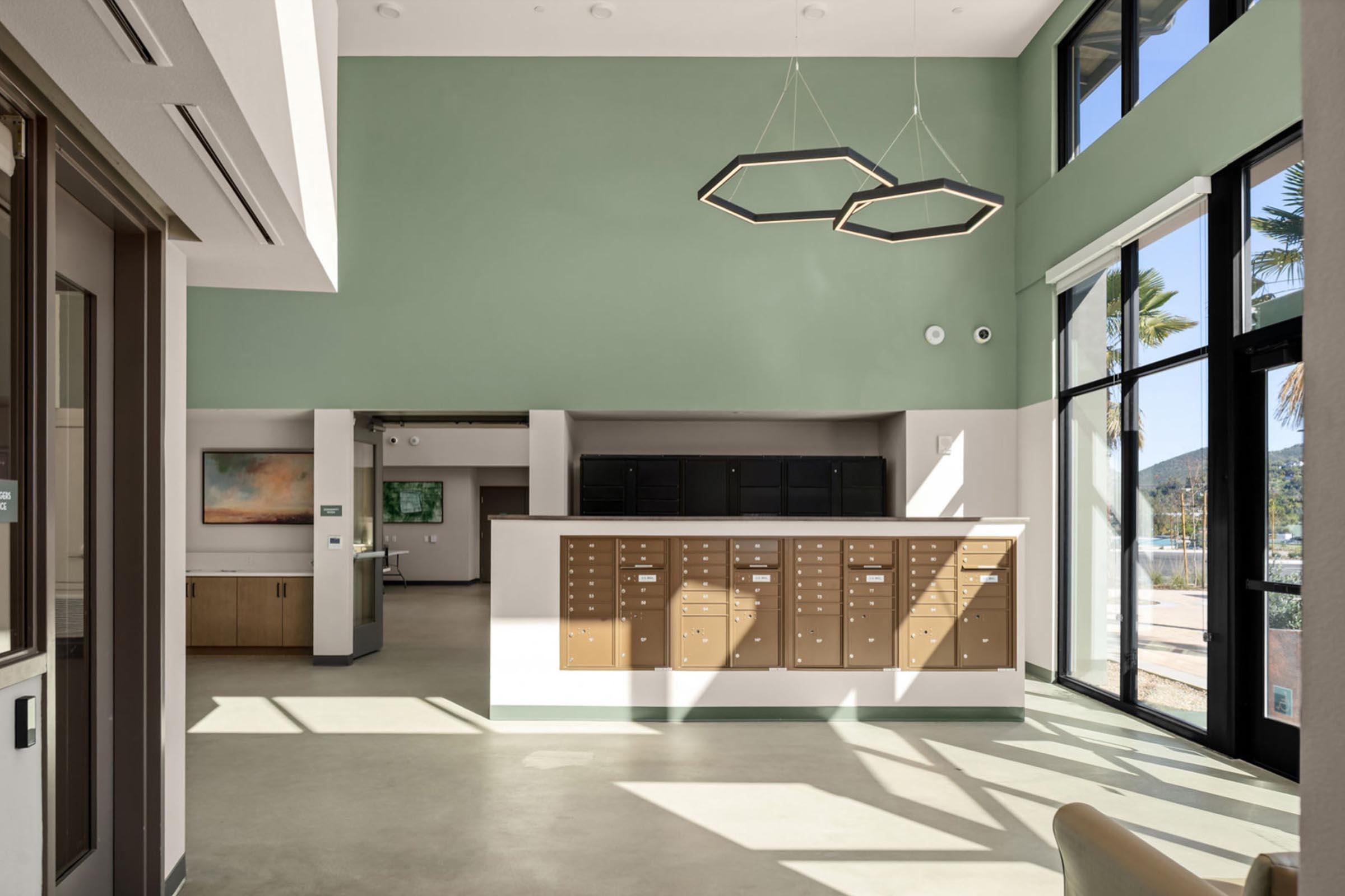 Bright and modern interior of a shared space featuring a green accent wall, large windows allowing natural light, a reception area with a counter, and mail lockers mounted on the wall. The design includes minimalistic lighting fixtures and a view of palm trees outside, creating an inviting atmosphere.