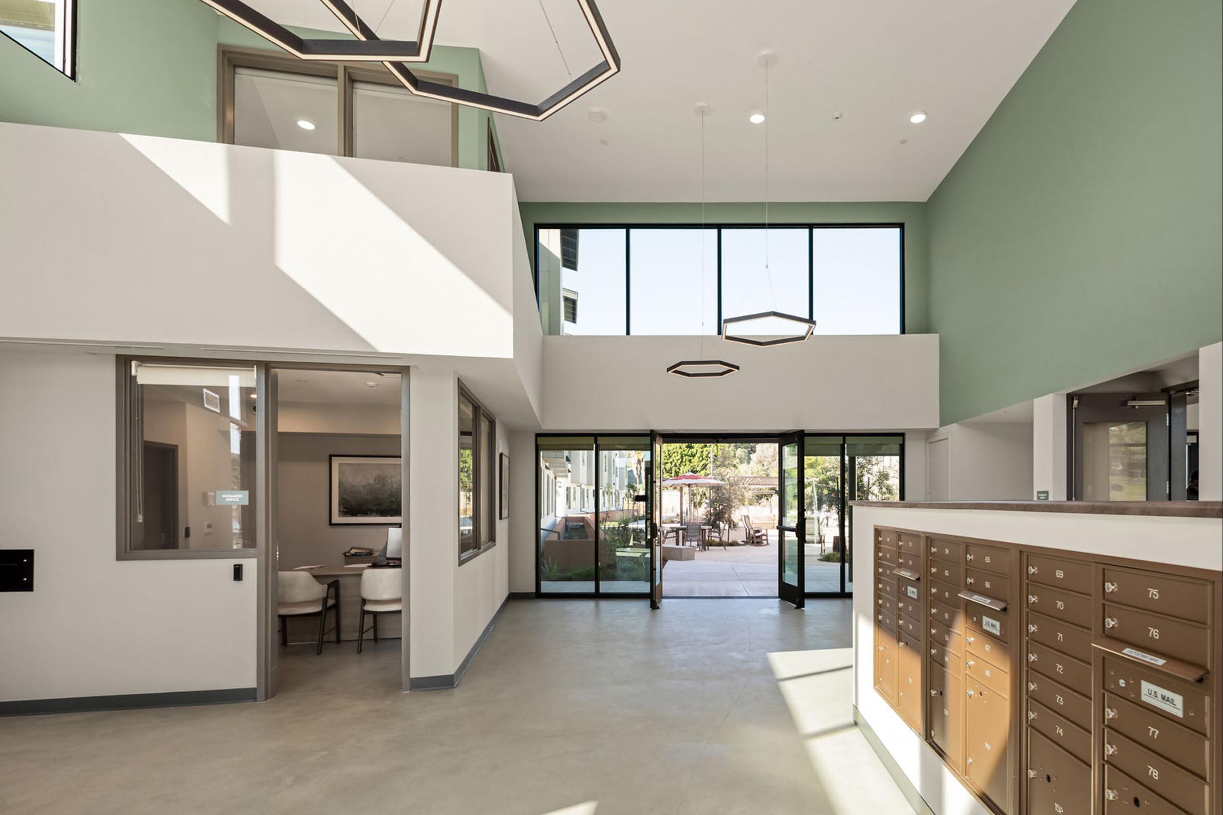 A spacious, modern lobby with large windows allowing natural light. The interior features a neutral color scheme with green accents, a seating area with circular tables, and a row of mailboxes on one wall. Outdoor views can be seen through the glass doors, leading to a patio area.