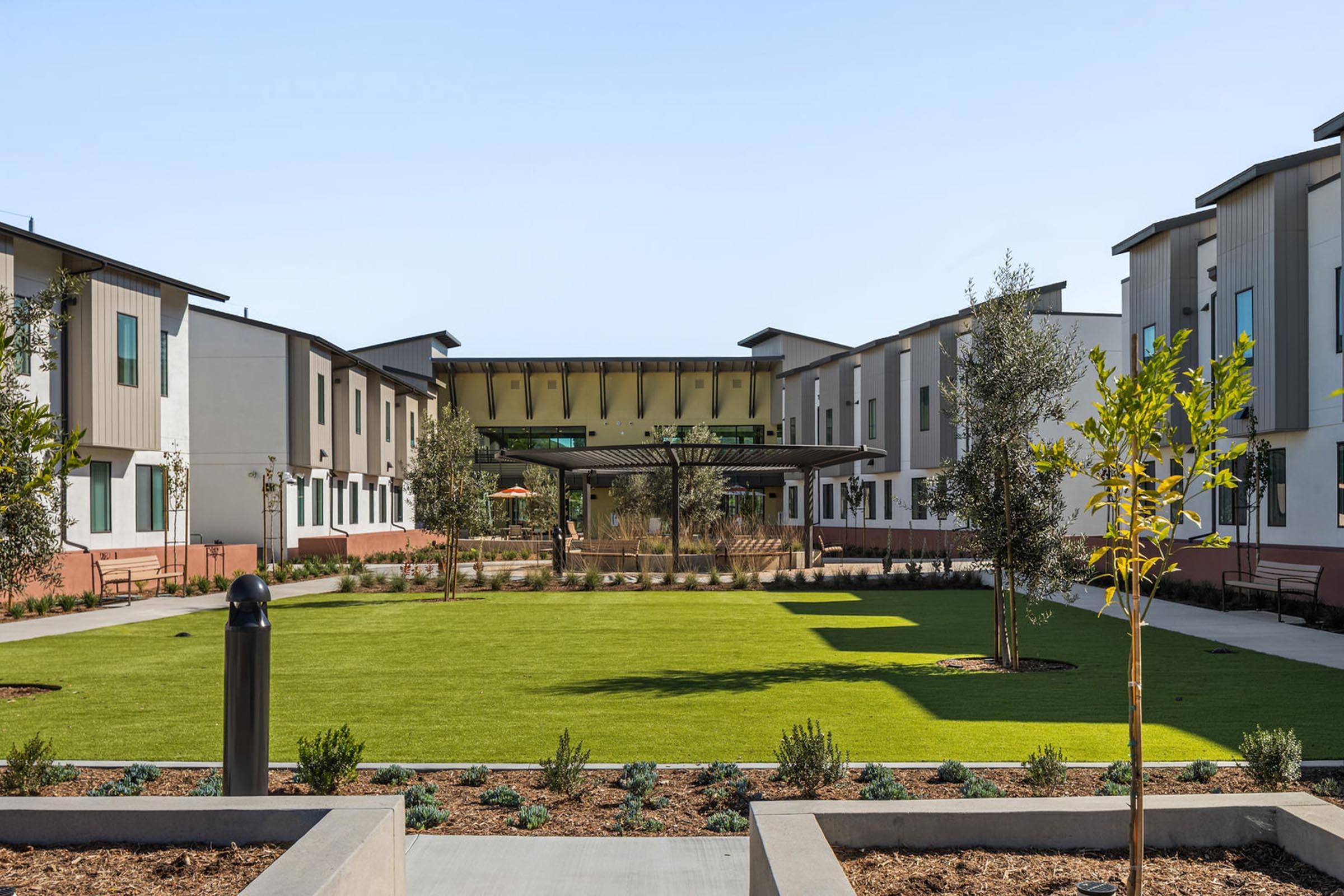 A modern residential complex featuring two-story buildings arranged around a green courtyard. The courtyard is landscaped with lawn and plantings, and there are benches and paths for walking. The sky is clear, creating a bright and welcoming ambiance.