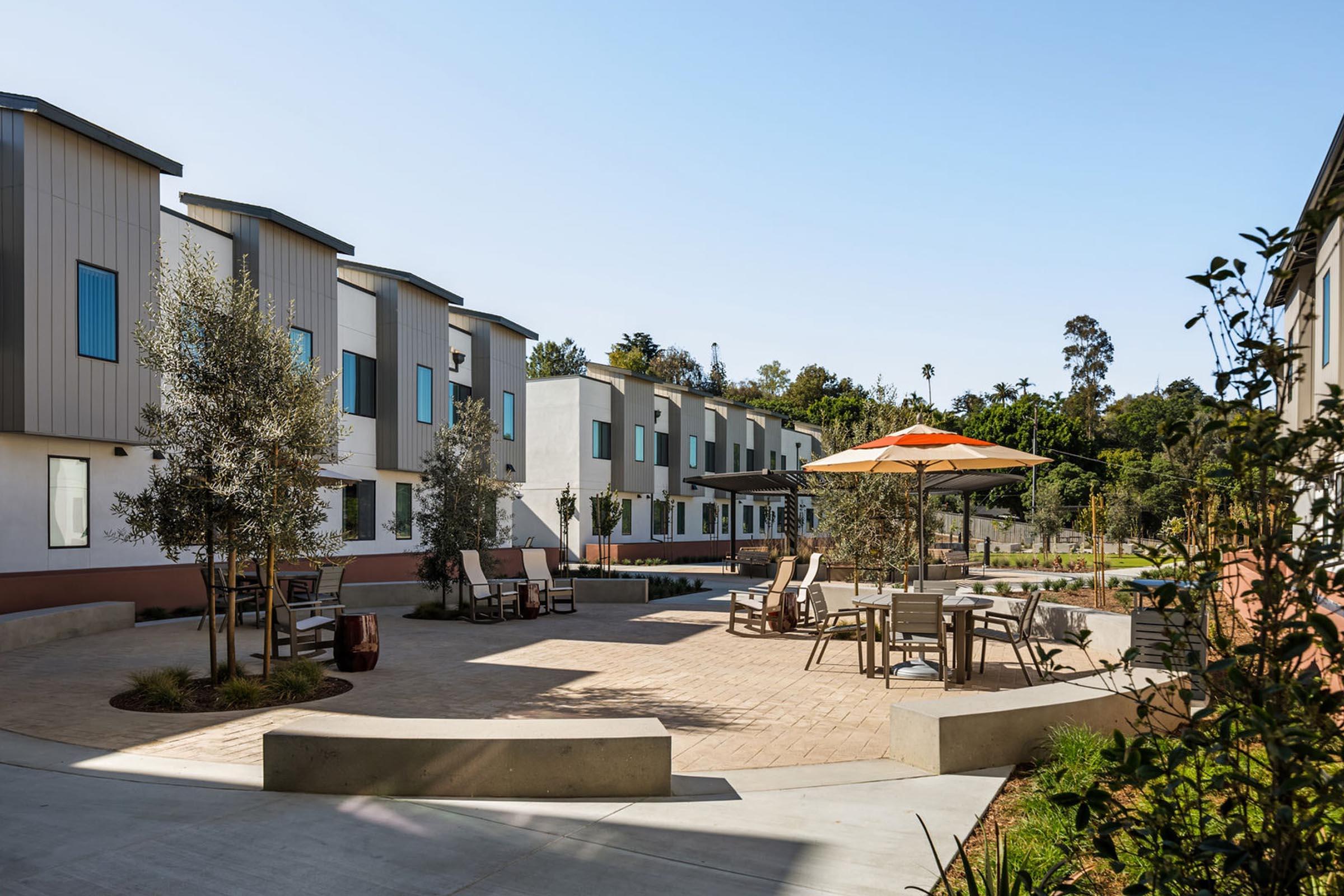 Modern residential complex featuring two-story buildings with large windows, surrounded by greenery. In the foreground, a landscaped courtyard with patio furniture and an orange umbrella provides a relaxing outdoor space. Clear blue sky enhances the bright and inviting atmosphere of the area.