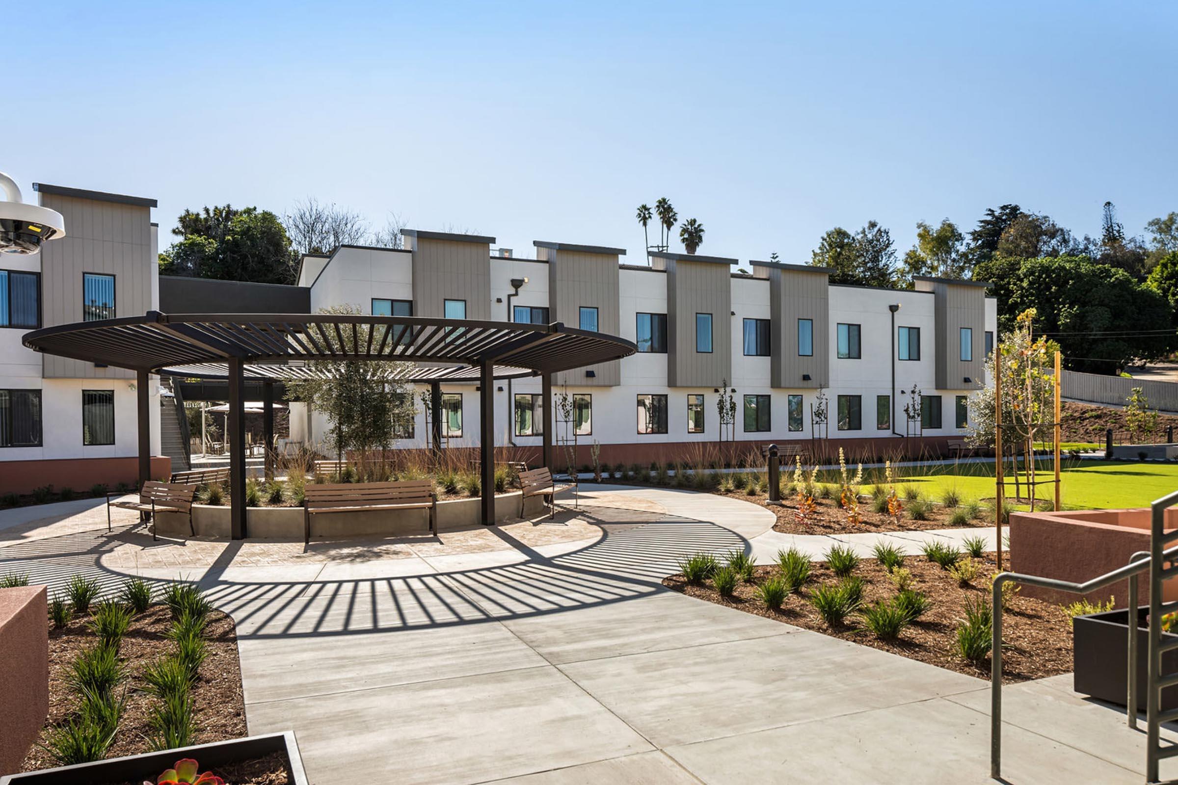A modern multi-unit building with landscaped gardens and walking paths. The area features a circular seating arrangement under a pergola, surrounded by greenery and palm trees. Blue skies and sunlight illuminate the scene, creating a welcoming outdoor space for residents.