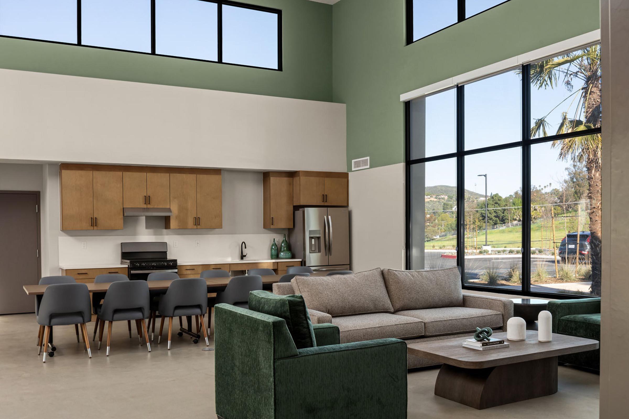 A spacious modern living area features a large gray sofa, green accent chairs, a wooden coffee table, and a sleek dining area with wooden cabinets and chairs. Floor-to-ceiling windows provide natural light and a view of the outdoors with palm trees and hills in the background.