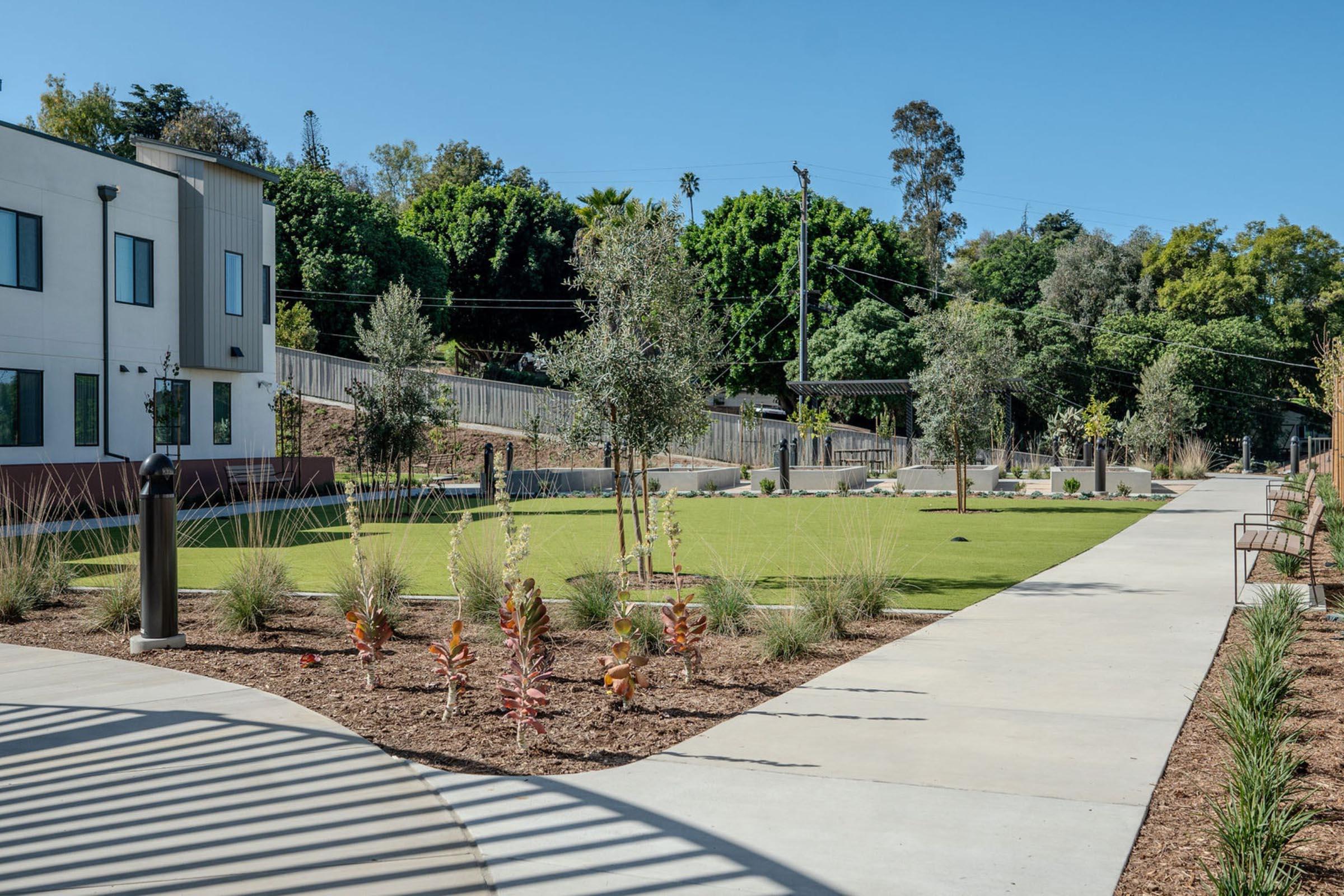 A landscaped park featuring a manicured lawn, young trees, and decorative shrubs. There are pathways through the greenery, with a modern building in the background and hills lined with trees. The scene is set against a clear blue sky, creating a serene outdoor environment.