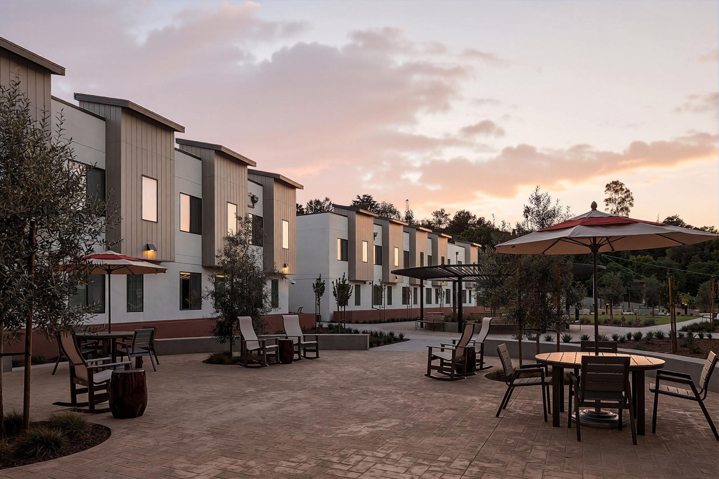 A modern residential complex at sunset, featuring several two-story buildings with large windows. In the foreground, a landscaped area includes a round wooden table, chairs, and umbrellas, surrounded by trees and shrubbery. The sky is painted in soft hues of pink and orange.