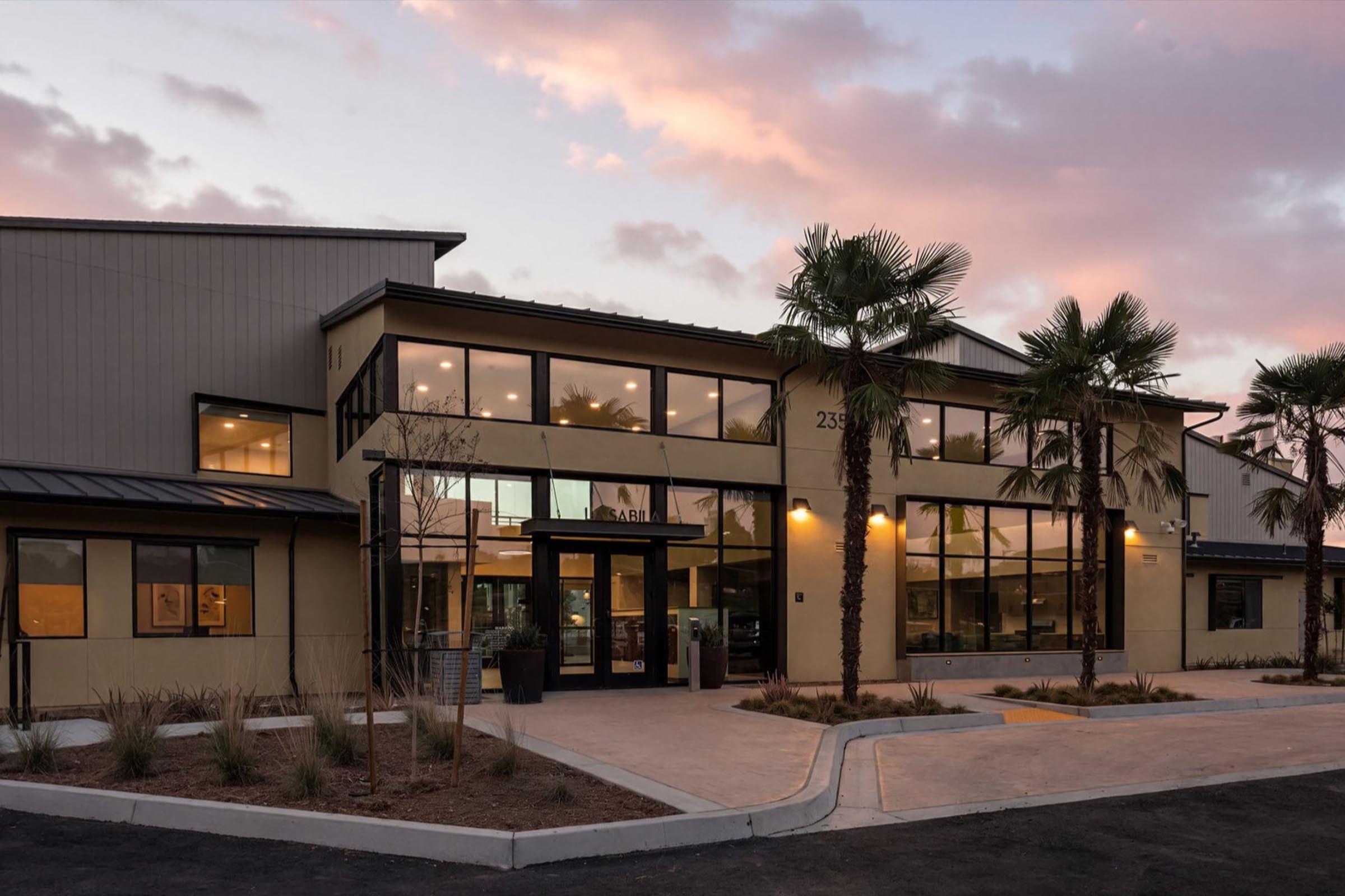 Modern building with large glass windows and a welcoming entrance, surrounded by palm trees. The structure features a mix of metal and stucco materials under a pastel sky at dusk, conveying a contemporary and inviting atmosphere. Driveway leads up to the entryway.