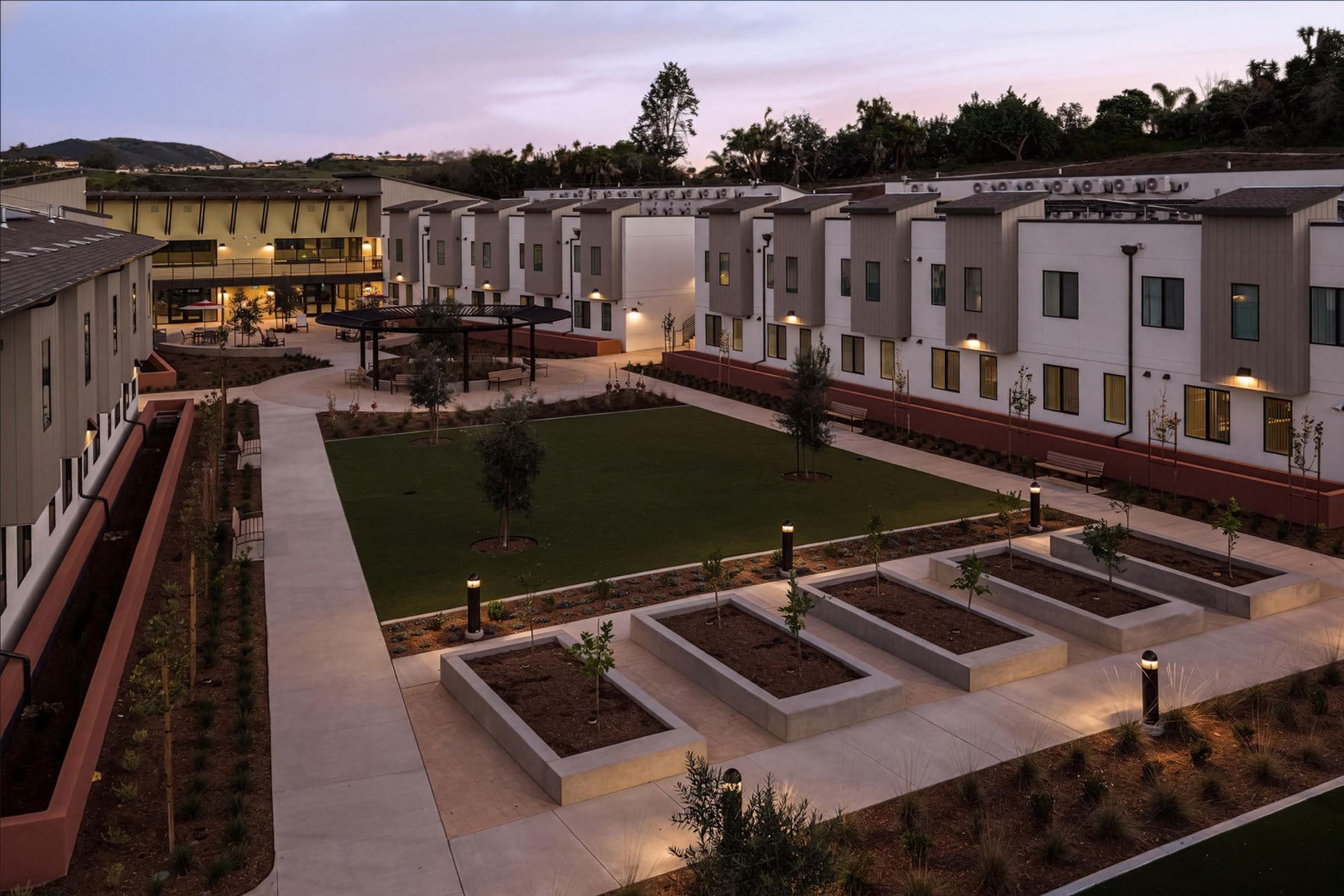 A modern residential courtyard featuring two-story buildings, greenery, and landscaped areas. Pathways connect the buildings, leading to an open green space with benches and raised garden beds. Soft outdoor lighting is installed along the paths, and the setting is illuminated by a twilight sky.