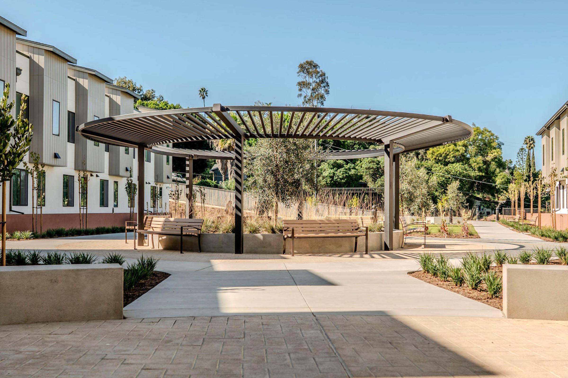 A modern outdoor space featuring a curved pergola with benches underneath, surrounded by neatly landscaped paths, plants, and residential buildings. Sunlight casts shadows on the pavement, creating a welcoming atmosphere for relaxation and social gatherings.