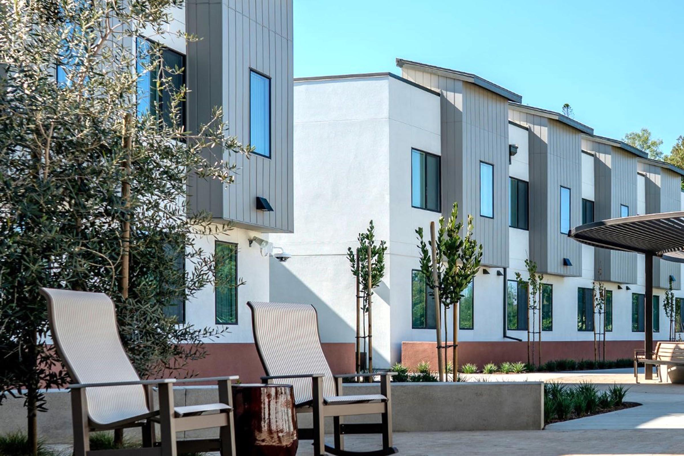 Modern multi-story building with a sleek exterior design, featuring clean lines and large windows. In the foreground, there are two lounge chairs and a small table surrounded by greenery. The scene is bright and well-lit, suggesting a welcoming outdoor space.