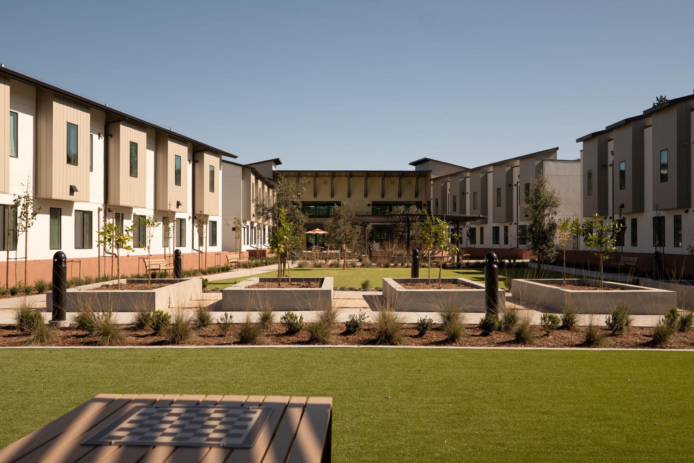 A view of a modern residential complex featuring two buildings flanking a landscaped courtyard. The courtyard has green grass, neatly arranged plants, and raised garden beds. A seating area with a table is visible in the foreground, under a clear blue sky.