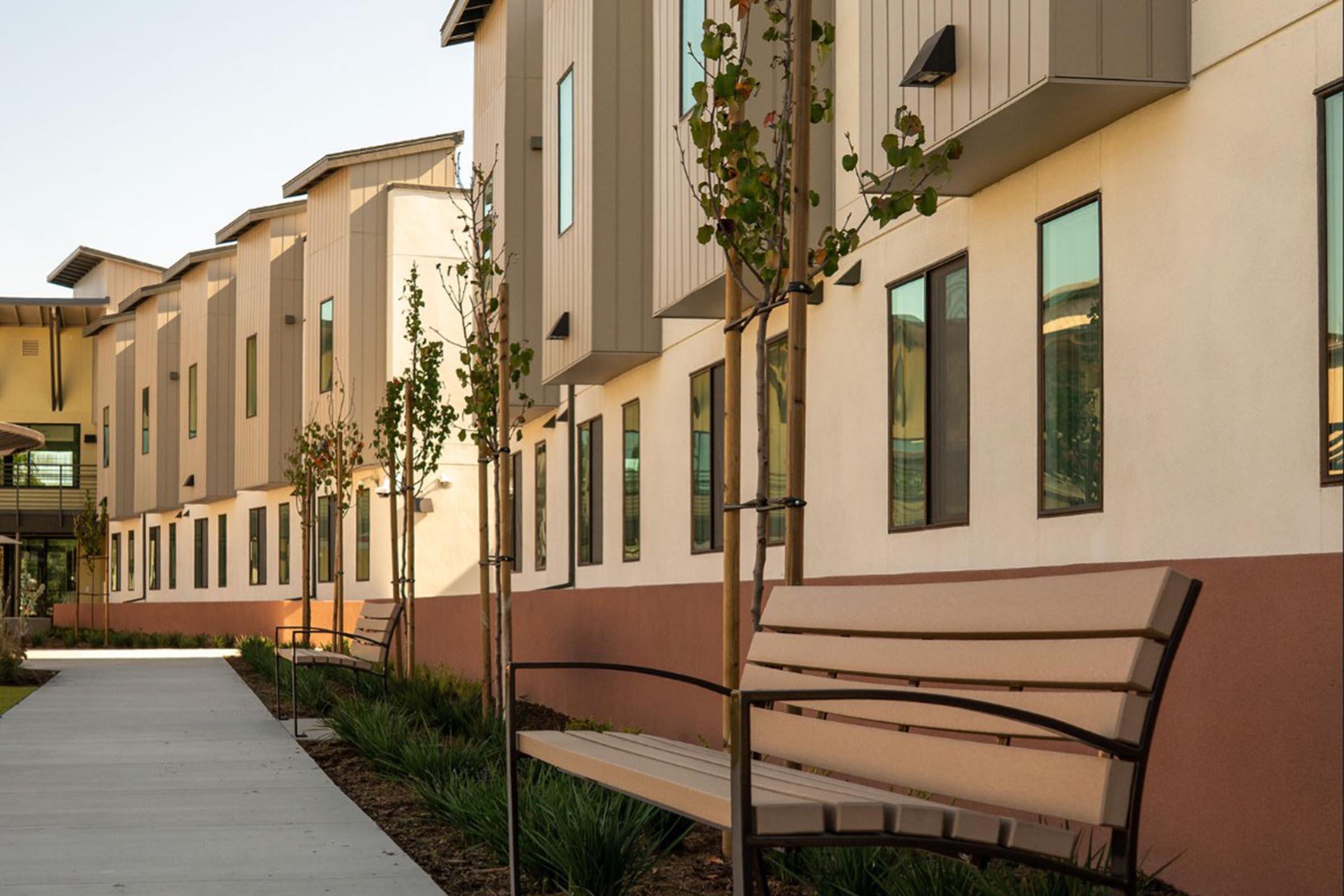 A modern building with a contemporary design featuring a series of windows and a smooth sidewalk in the foreground. Young trees line the path, and a bench is positioned along the walkway, creating a welcoming outdoor space. The overall atmosphere is clean and inviting.