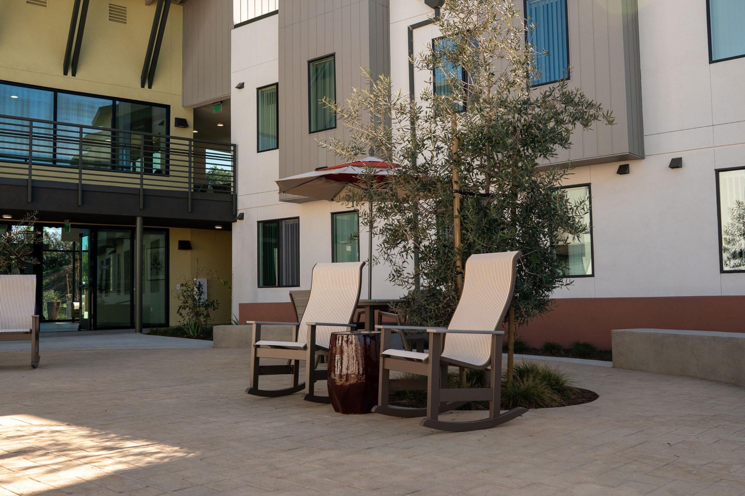 Two rocking chairs with a small side table are positioned under a red umbrella in a courtyard. The area features light-colored paving stones and greenery, including shrubs and a tree. In the background, there are modern buildings with large windows, creating a relaxing outdoor space.