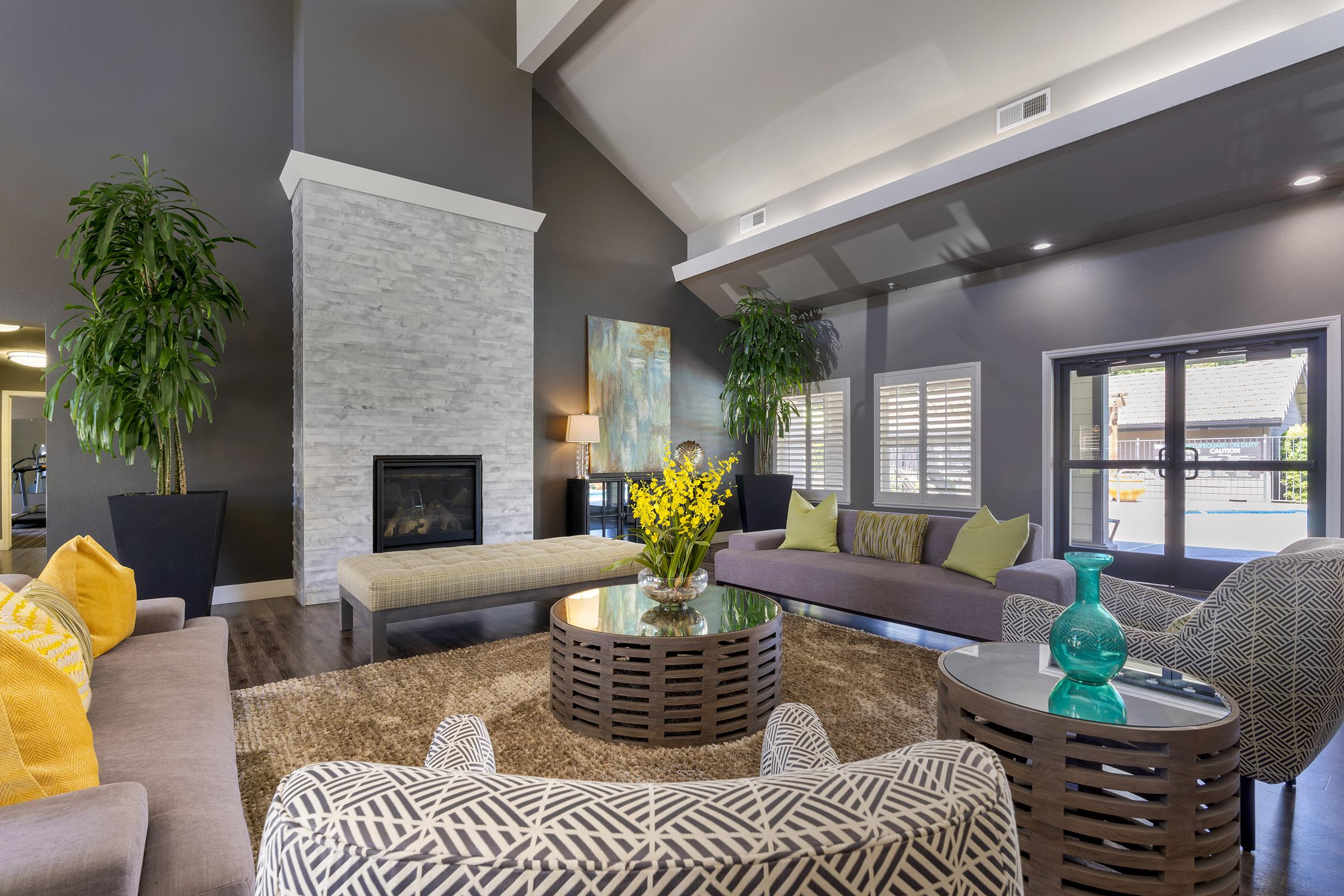 A modern lounge room featuring a high ceiling, stone fireplace, and large windows. The space is furnished with a circular coffee table surrounded by sofas in shades of gray and yellow cushions. Decorative plants and a vibrant piece of artwork add a stylish touch. Natural light brightens the cozy atmosphere.