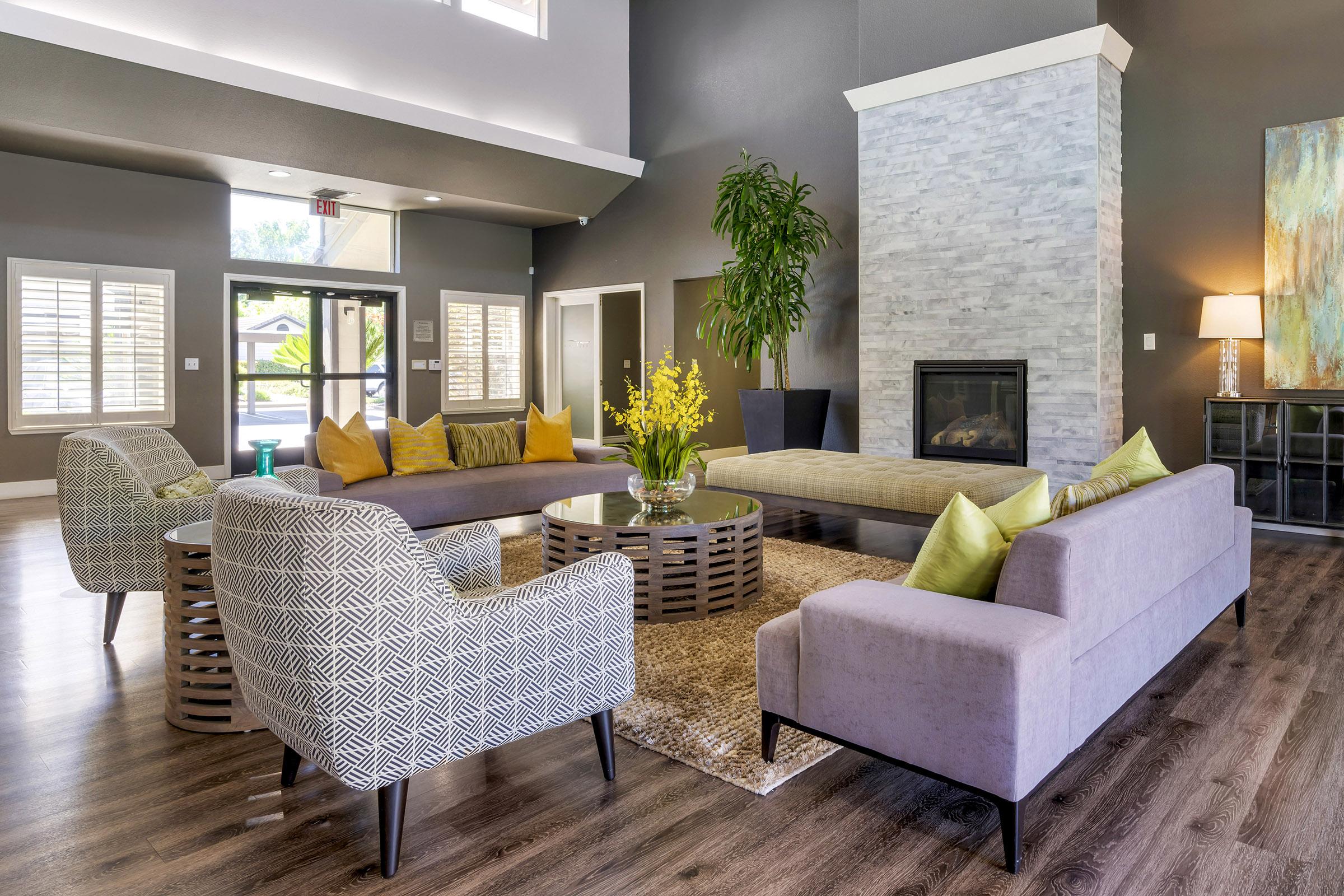 Modern lounge area featuring a circular coffee table surrounded by gray sofas and patterned armchairs. There are yellow decorative pillows, a plant in the corner, and a stone fireplace. Large windows provide natural light, creating a welcoming atmosphere with neutral-toned walls and hardwood flooring.