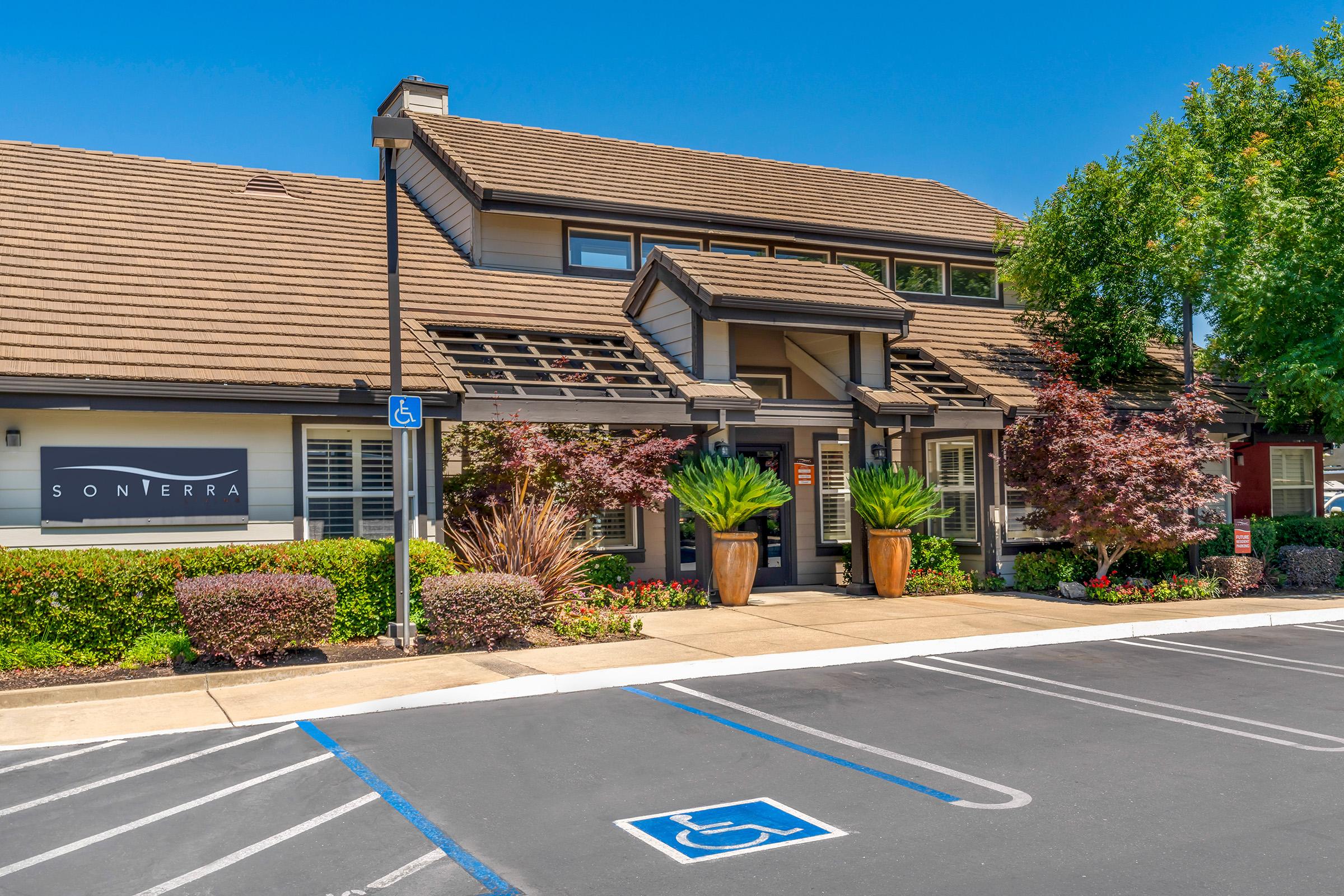 A modern building with a sloped roof, featuring a sign for "Sonterra." The exterior is landscaped with colorful plants and trees. There are large pots near the entrance and designated parking spaces, including a handicap spot, in front of the building. The sky is clear and blue, indicating a sunny day.
