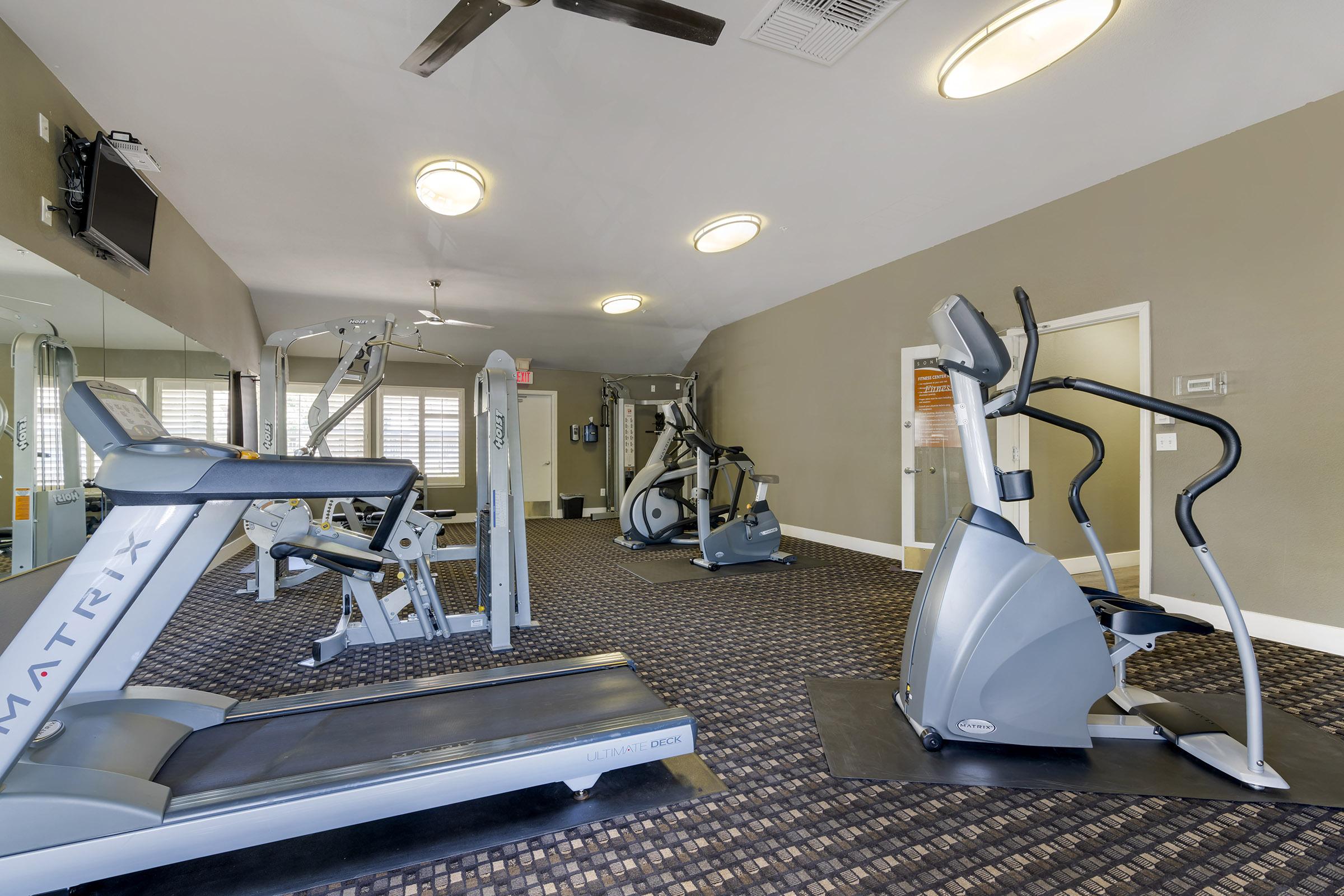 A modern gym interior featuring exercise equipment including a treadmill, elliptical machine, and weight machines. The space is well-lit with ceiling fans and wall mirrors, providing a clean and inviting atmosphere for workouts.