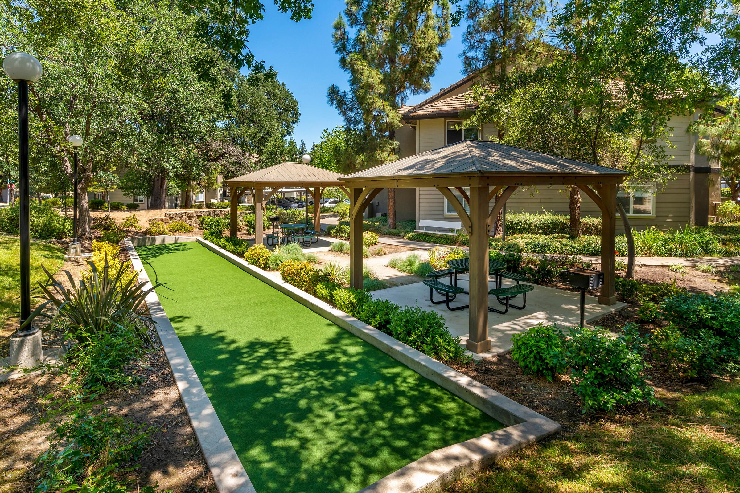A landscaped area featuring two wooden gazebos with picnic tables, surrounded by green grass and garden plants. A bocce ball court runs between the gazebos, all set in a bright, sunny environment with trees in the background.