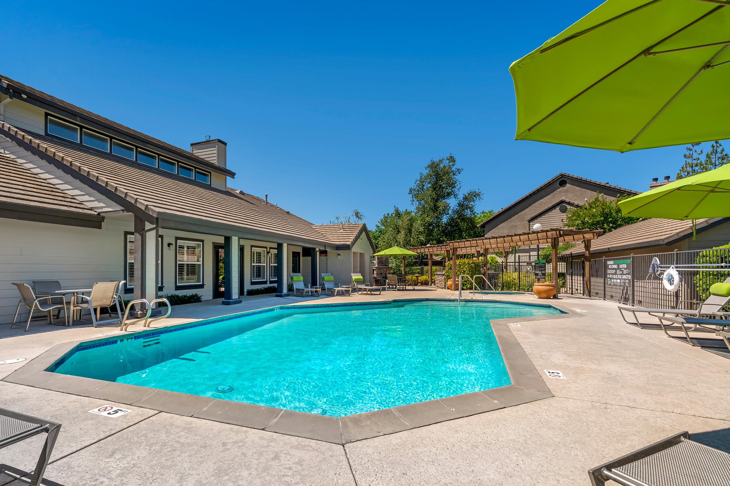 A vibrant outdoor swimming pool surrounded by green umbrellas and lounge chairs. The pool area features a clean, inviting atmosphere with well-maintained landscaping and a building nearby. Sunlight shines down, creating a refreshing vibe ideal for relaxation and leisure.