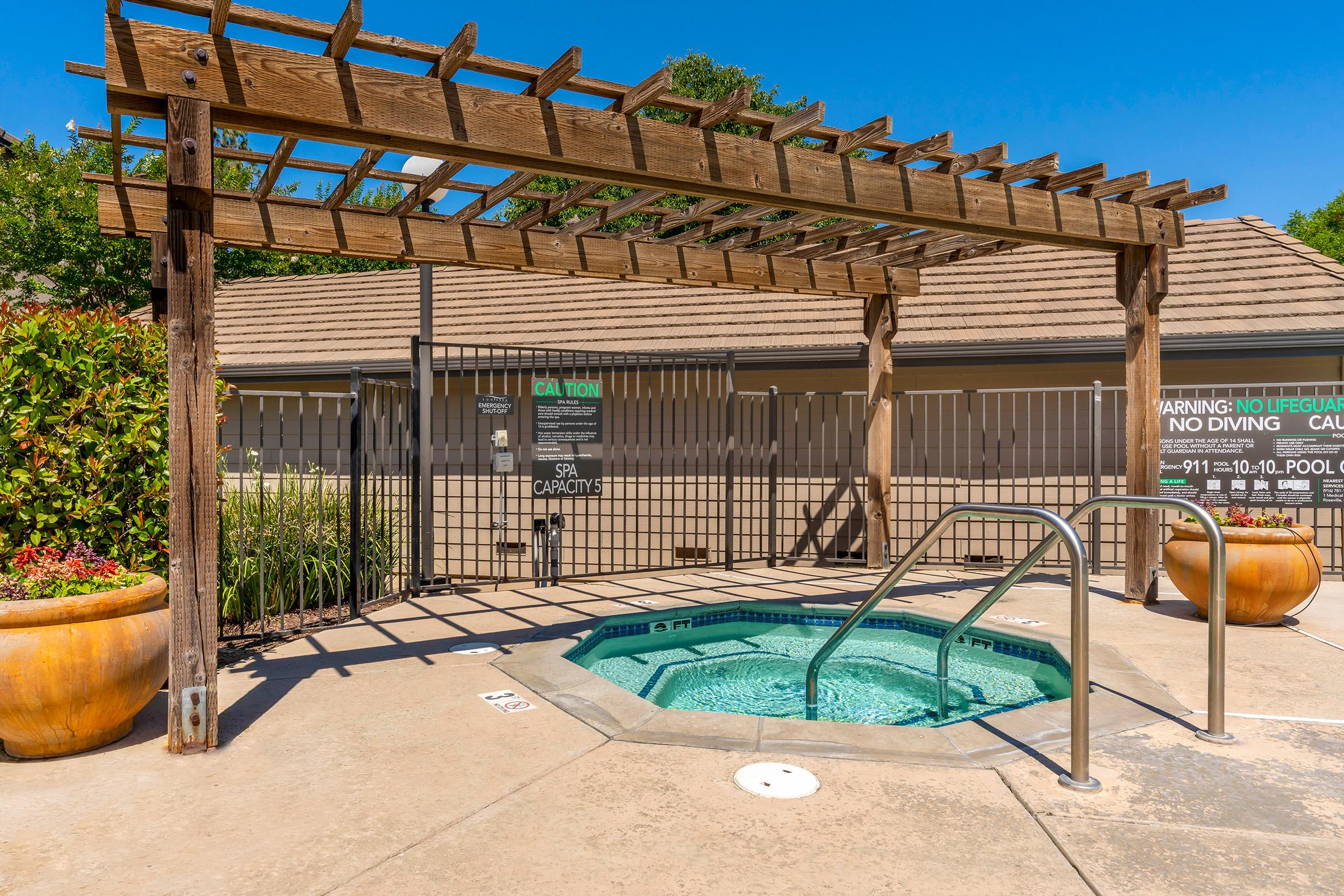 A small outdoor spa or hot tub located under a wooden pergola. The area is surrounded by a fence and features two large planters. There are warning signs about pool safety visible, with clear blue skies in the background. The overall setting is clean and well-maintained.