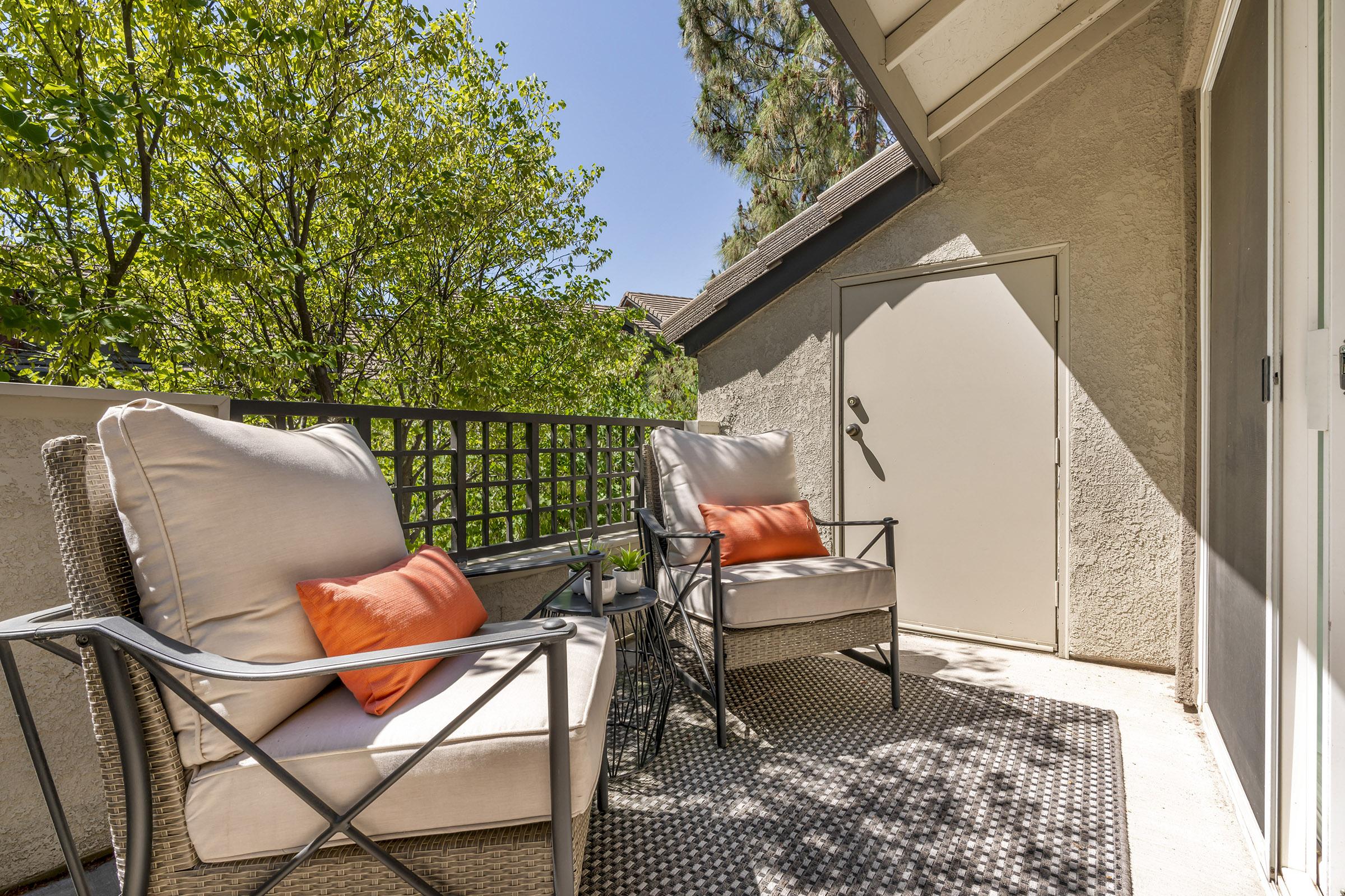 A cozy outdoor balcony featuring two cushioned chairs with orange pillows, a small table between them, and surrounded by greenery. The flooring is patterned, and a door is visible in the background, adding to the inviting atmosphere. Bright sunlight illuminates the space, creating a warm and welcoming setting.